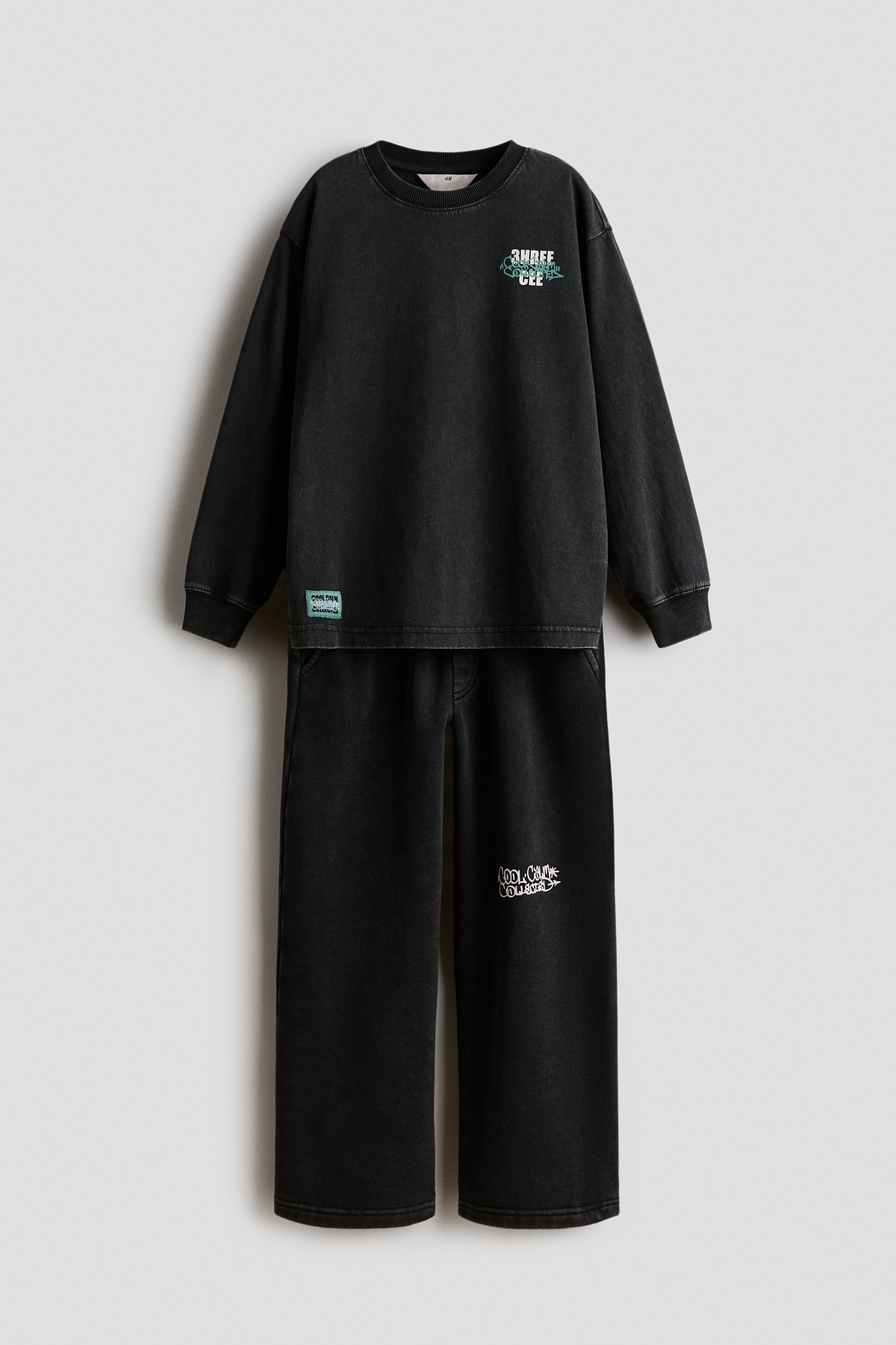 A child is wearing a front-facing, dark green matching set consisting of a long-sleeve jersey top and straight-leg track pants. The top features a ribbed crew neckline, ribbed cuffs, and a small white printed design on the chest, styled over a layered white T-shirt. The matching track pants feature side pockets and a white graphic print on the left thigh.