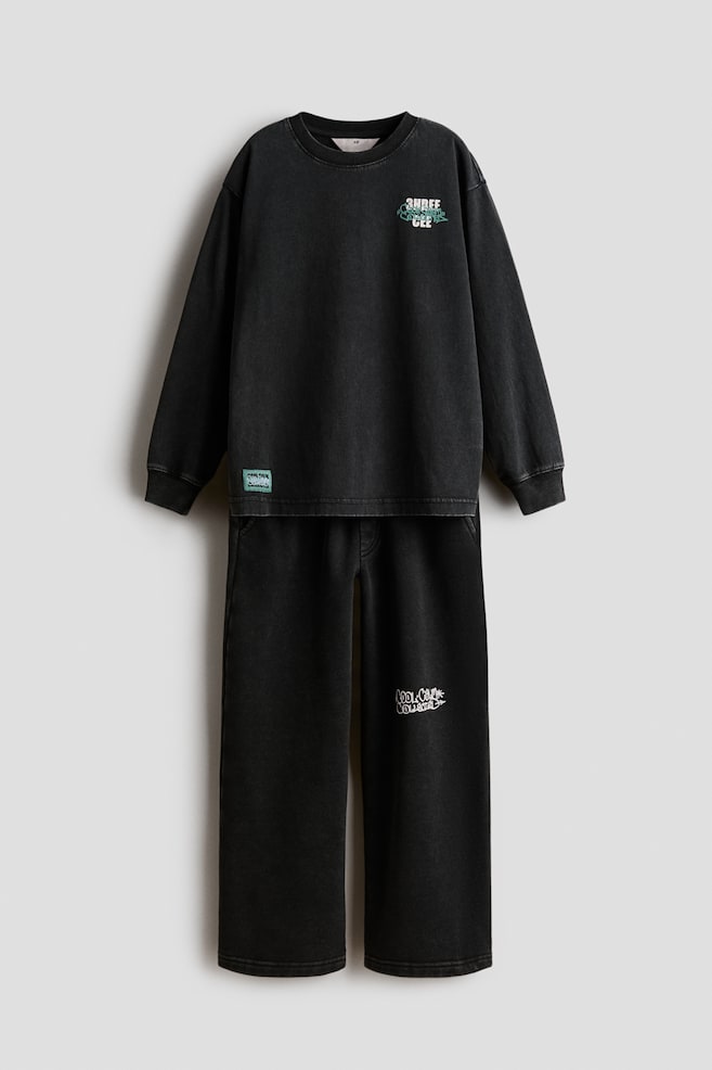 Cet ensemble assorti deux pièces à fini noir délavé comprend un sweat-shirt à manches longues et un pantalon à jambe droite, présenté de face. Le sweat-shirt met en valeur un col rond côtelé et des poignets côtelés, un petit graphique sur le haut de la poitrine gauche et une petite écusson vert près de lâourlet, tandis que le pantalon coordonné affiche une coupe décontractée avec un petit graphique sur la cuisse gauche. - 1