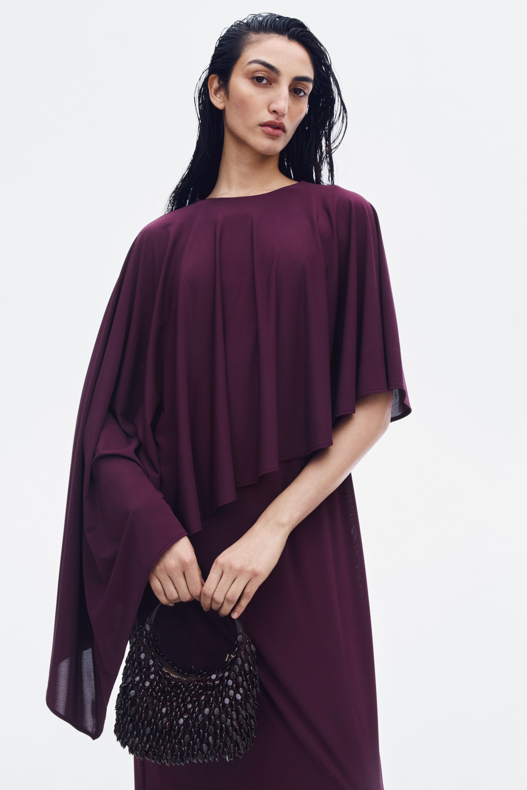 View larger image: A dark purple jersey dress featuring a round neckline and a draped, asymmetrical cape-like overlay is being worn by the model. The model is holding a small, dark, beaded handbag in front of the dress.