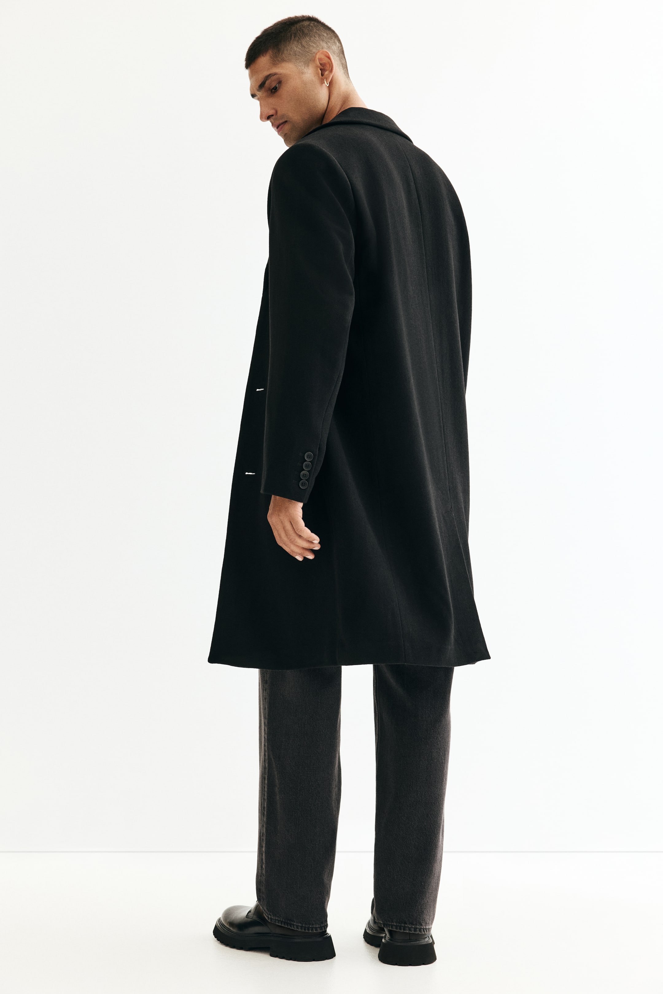 View larger image: A black coat is being shown from a back-to-side angle. It is featuring notch lapels and a single-breasted button closure. The long coat is extending past the hips, and four decorative buttons are visible on the right sleeve cuff.