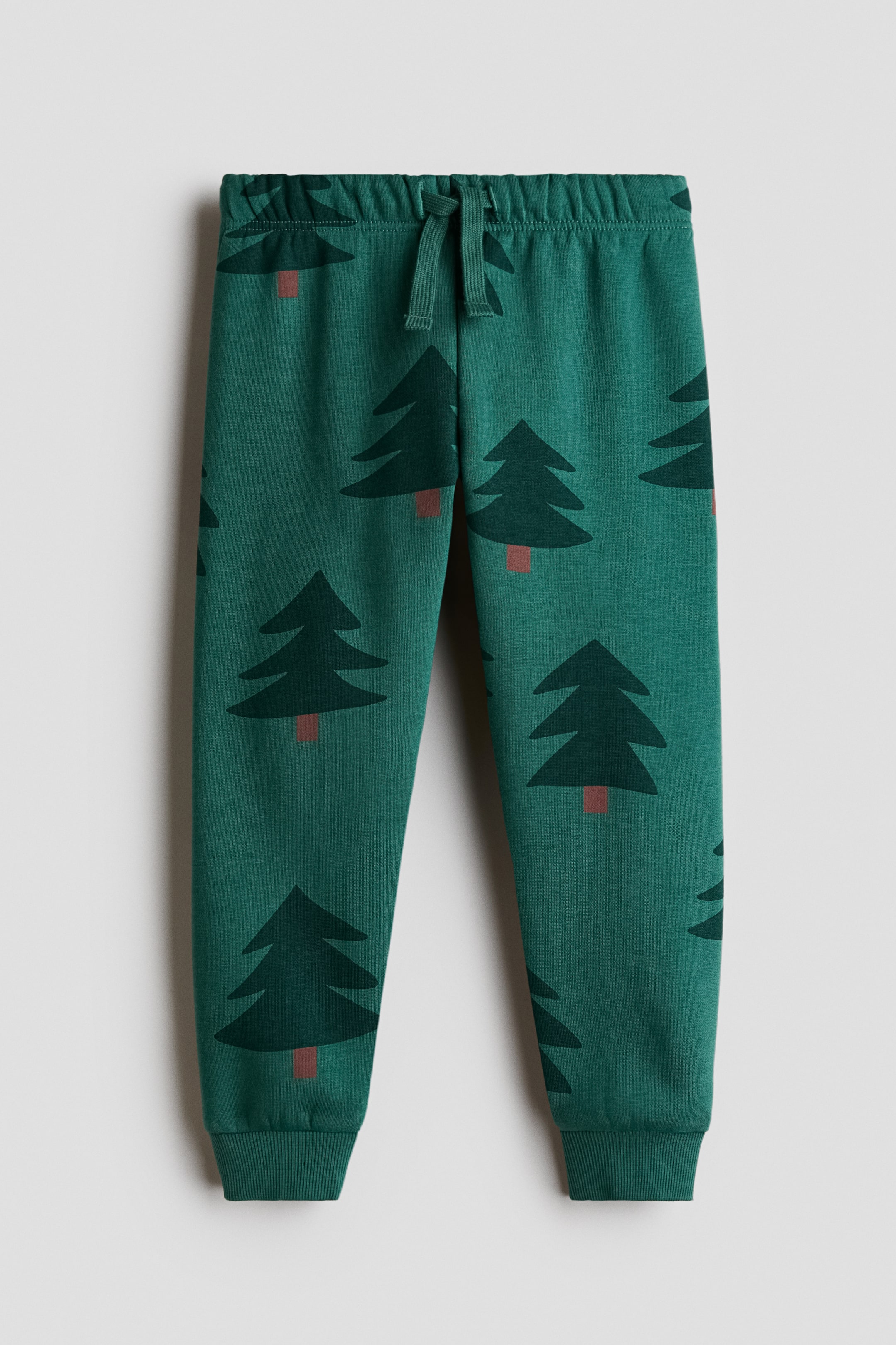 View larger image: These green sweatpants feature an all-over print of dark green pine trees with brown trunks, shown from the front. The design includes an elastic waistband with a matching drawstring and ribbed ankle cuffs.