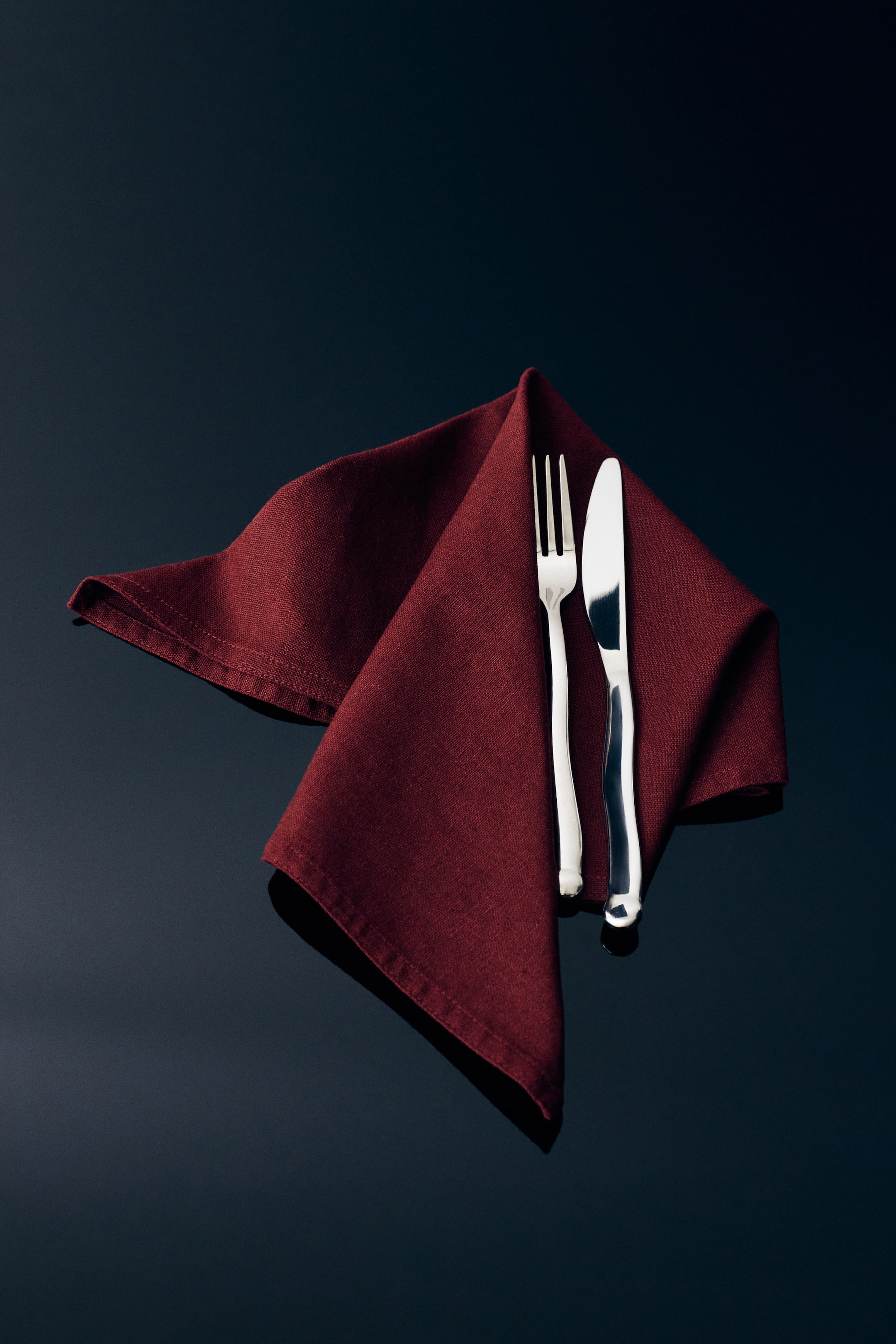 View larger image: A dark red napkin in a textured linen and cotton blend displays stitched hems while being folded artistically. A silver fork and knife rest on the right fold of the napkin.