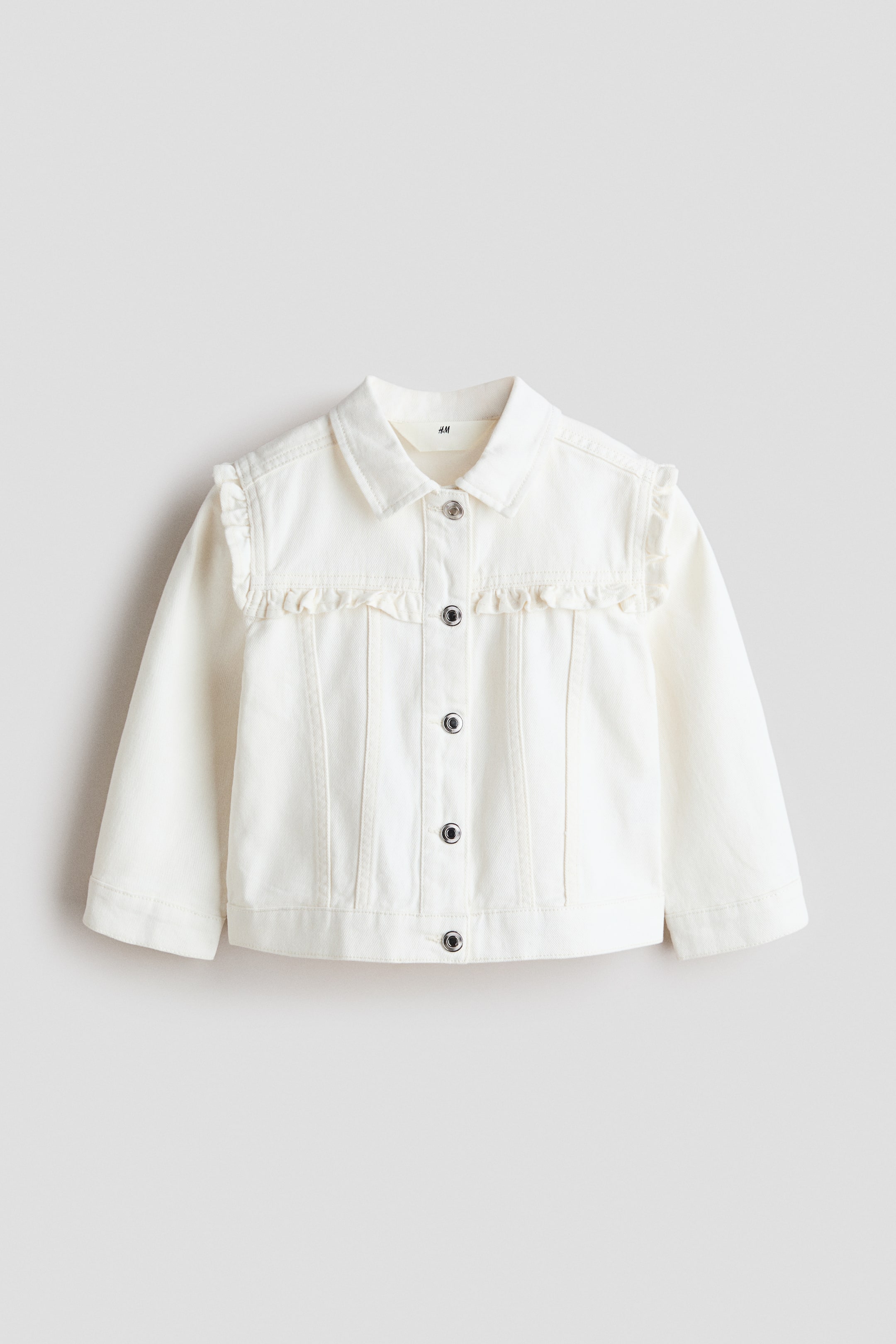 View larger image: A white denim jacket faces forward, with a standard collar and silver-tone snap buttons down the center. Ruffles adorn the front yokes and extend over the shoulders, with vertical seams defining the front panels.
