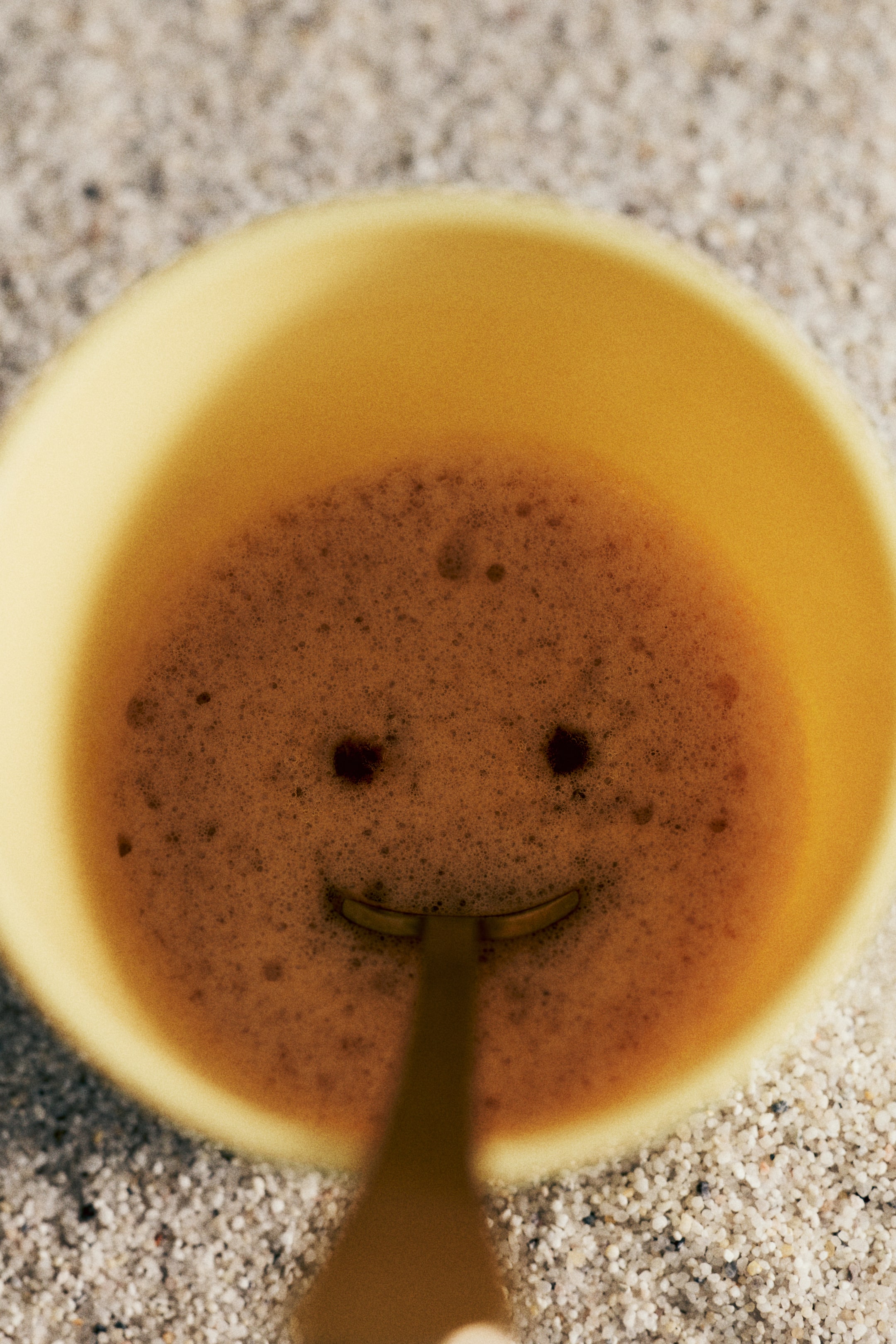 Visualizza immagine più grande: Una tazza gialla contiene un liquido marrone e schiumoso. La schiuma forma un volto sorridente con due macchie scure e rotonde per gli occhi, mentre la punta di un cucchiaio crea la bocca curva.