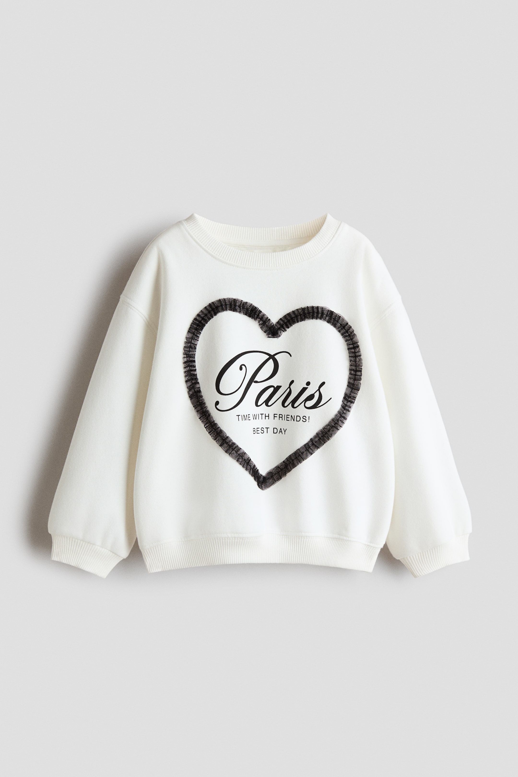 Se større billede: En cremefarvet sweatshirt med rund hals vender forfra og har sænkede skuldre, ribkanter ved ærmerne og en ribkant forneden. På brystet ses en stor hjerteformet grafik med en mørk, bølget kant, som indeholder den sorte skrifttype “Paris” over den trykte tekst “TIME WITH FRIENDS!” og “BEST DAY.”