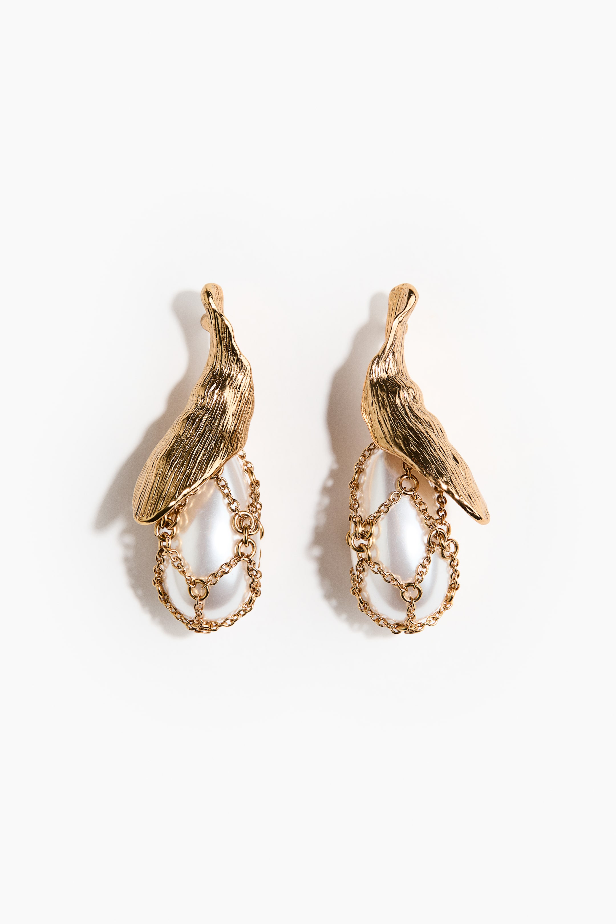 View larger image: A pair of earrings is showcasing a textured, organic gold-toned top component. An elongated, iridescent pearl is dangling from this component, being encased by a delicate gold-toned chain mesh.