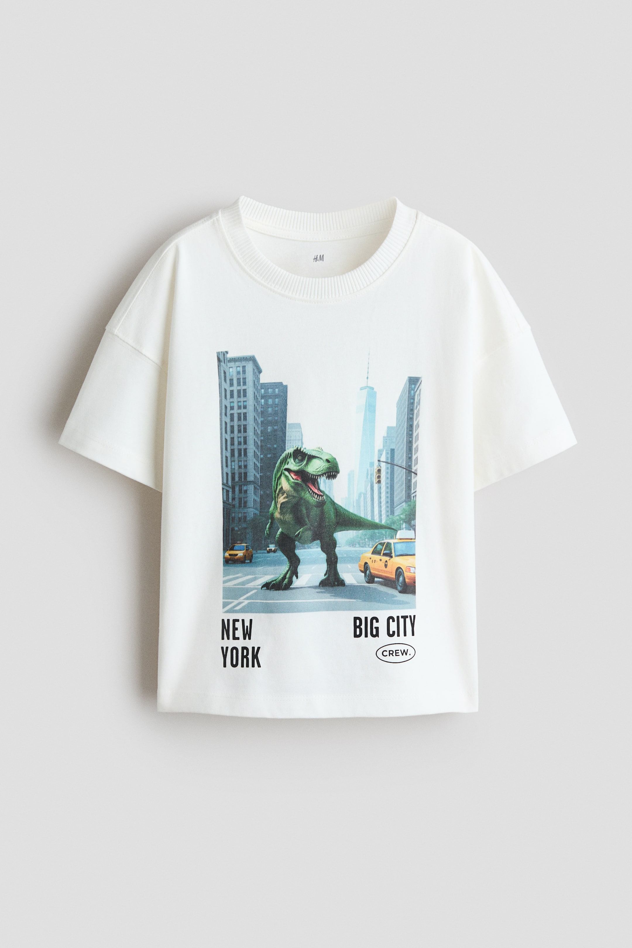 View larger image: A cream-colored t-shirt showcases a large graphic print on its front, featuring a green Tyrannosaurus Rex dinosaur standing on a city street with tall buildings and yellow taxi cabs. The t-shirt is styled with a ribbed crew neck and short sleeves, and the print includes the text "NEW YORK" and "BIG CITY CREW."
