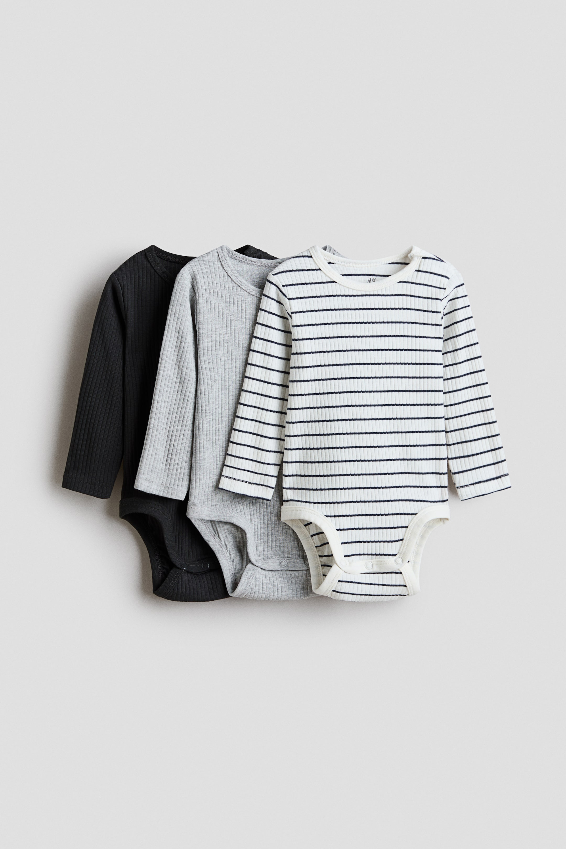 View larger image: Three long-sleeved bodysuits are showcased in soft, ribbed cotton jersey, transitioning from a black one, to a light grey one, to a white one with black horizontal stripes. Each bodysuit features a round neck, a concealed press-stud on one shoulder, and discreet press-studs at the bottom.
