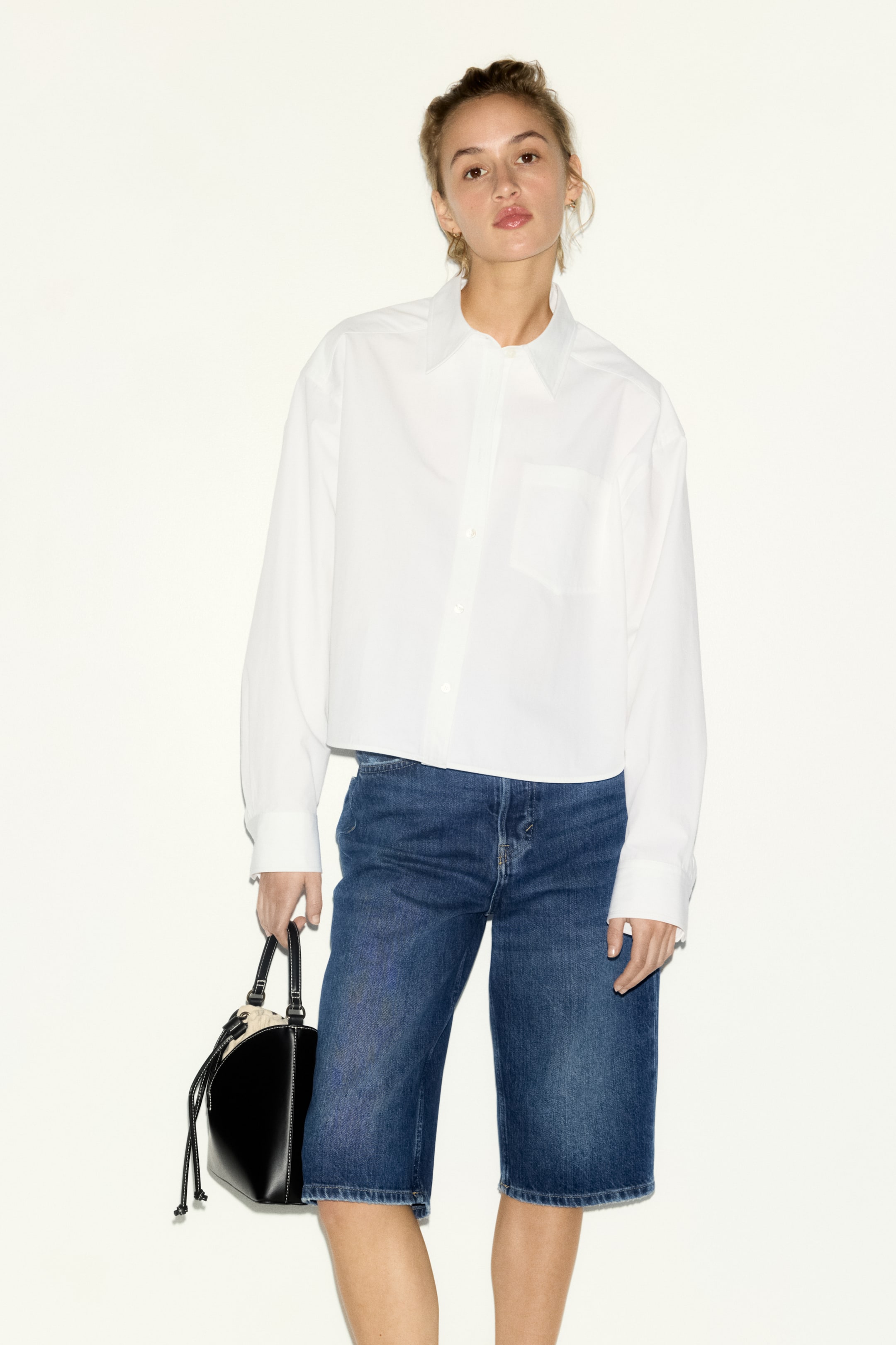 이미지 확대 보기: 앞면을 향해 있는 흰색 크롭 셔츠가 소개되고 있습니다. 이 셔츠는 뾰족한 칼라, 버튼 프론트 클로저, 그리고 버튼 커프스가 있는 긴 소매가 특징입니다. 왼쪽 가슴에 단일 패치 포켓이 보이며, 파란색 데님 버뮤다 반바지 위에 스타일링되어 편안하고 크롭된 핏을 강조하고 있습니다.