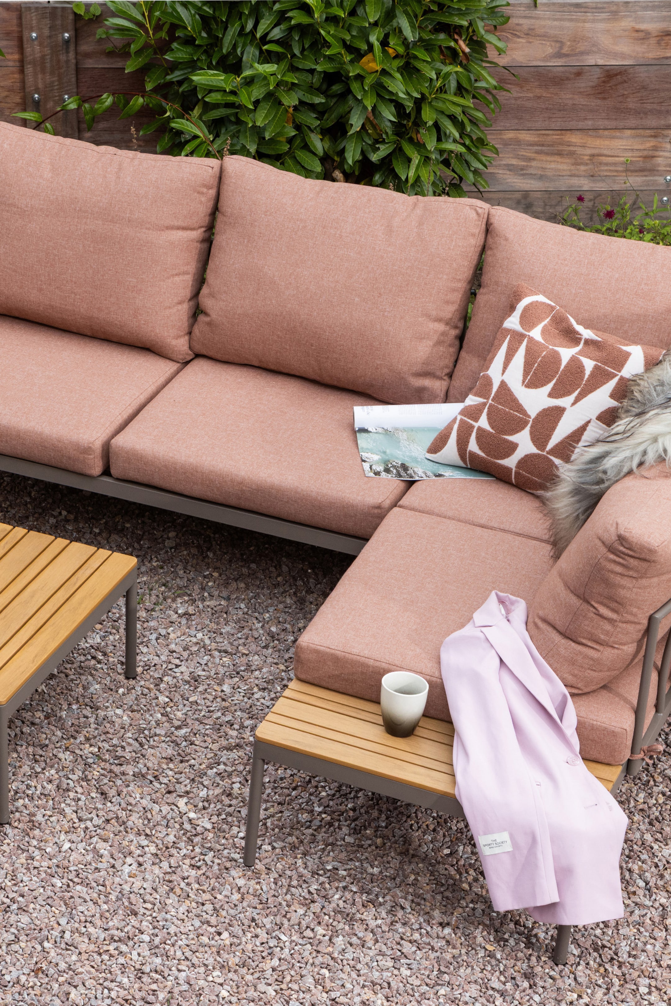 Agrandir l'image: Un canapé d'angle d'extérieur présente des coussins en tissu rose saumon reposant sur un cadre en métal gris foncé. Le canapé forme un L et intègre un accoudoir en bois à lattes sur son côté droit.