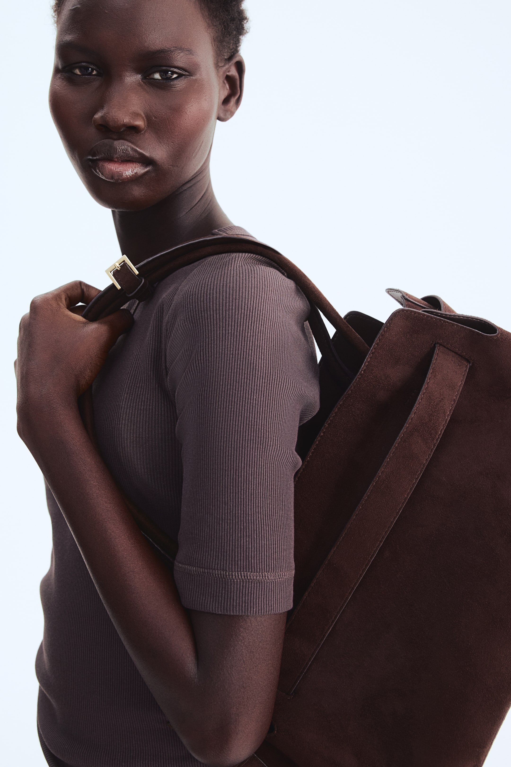 Zobrazit větší obrázek: Hnědá semišová taška je nesena přes rameno, její hlavní tmavě hnědý popruh se zlatým sponou spočívá na rameni. Taška se vyznačuje měkkou, neformální siluetou a kratší tmavě hnědé rukojeti jsou viditelné uvnitř otevření tašky.