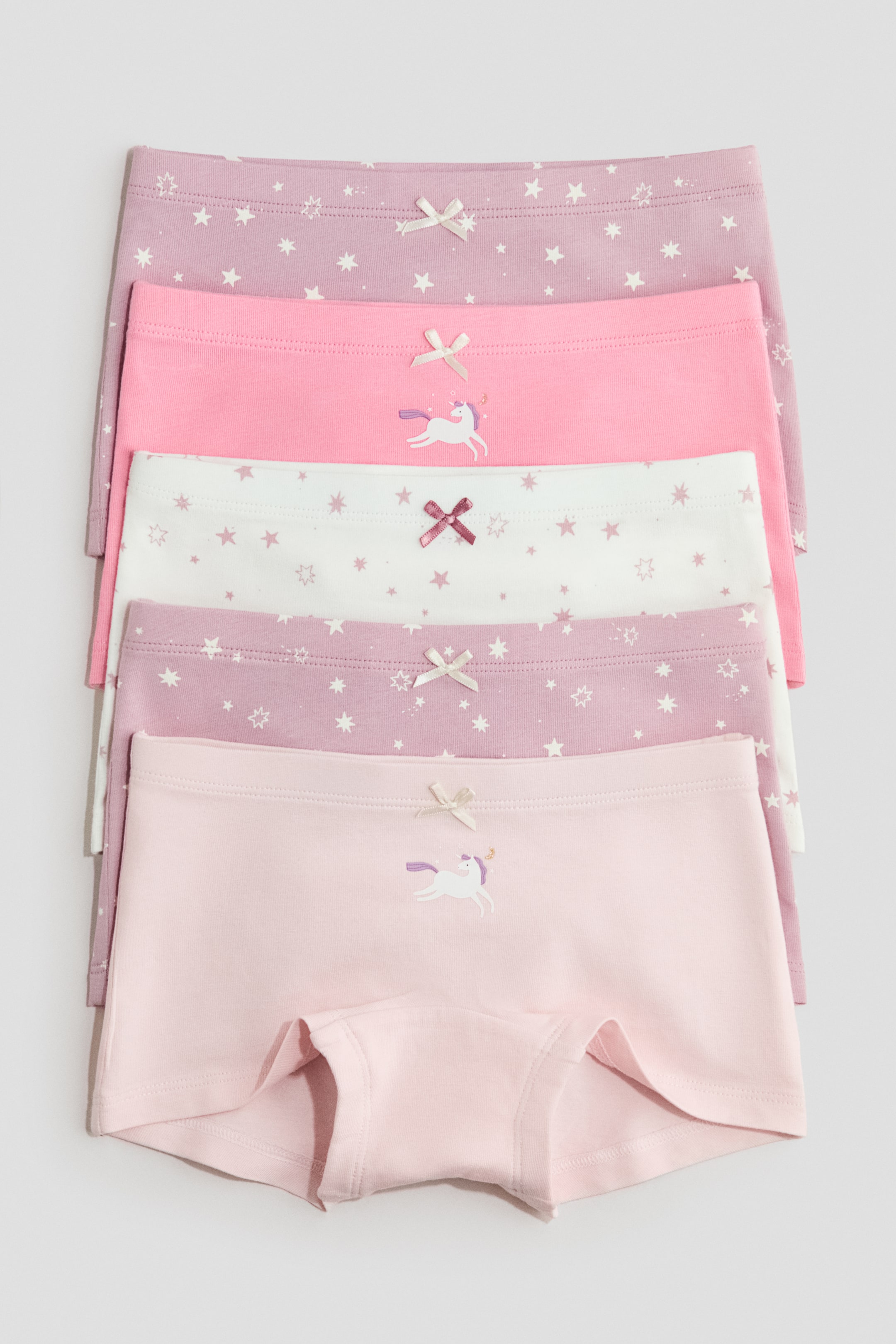 View larger image: A set of five children's boxer briefs is arranged, with their front sides facing forward. The briefs showcase varying shades of pink, white, and light purple, with some featuring star patterns or a unicorn design. Small decorative bows adorn the waistbands.
