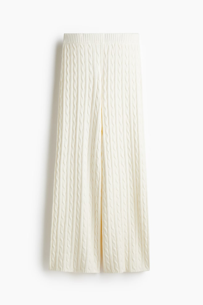 Pantalones de punto trenzado color crema se muestran extendidos, con una cintura acanalada, piernas anchas y patrones de punto trenzado vertical que cubren toda la superficie. - 1