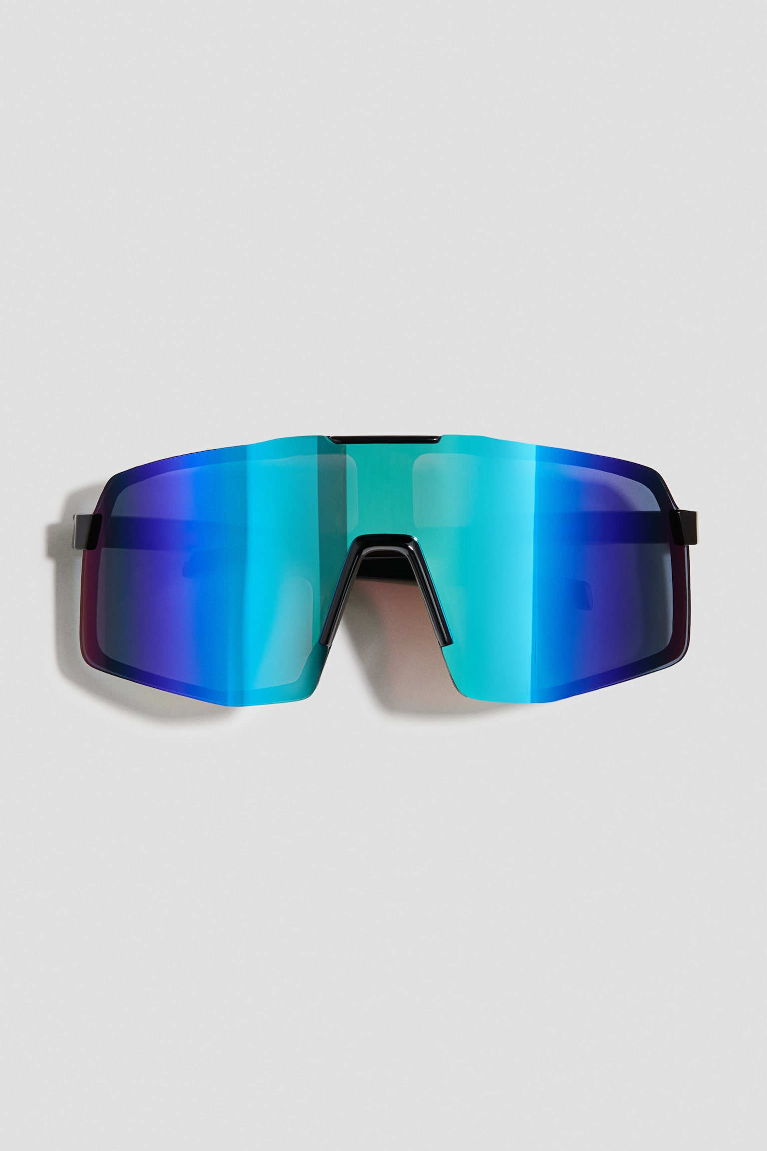 Die von vorne ausgerichtete Sonnenbrille verfÃ¼gt Ã¼ber eine einteilige, Ã¼bergrosse Shield-Glasfront mit einem lebendigen verspiegelten Farbverlauf in Blau, GrÃ¼n und Violett. Die Sonnenbrille zeigt einen schwarzen Rahmen an Steg und BÃ¼geln.