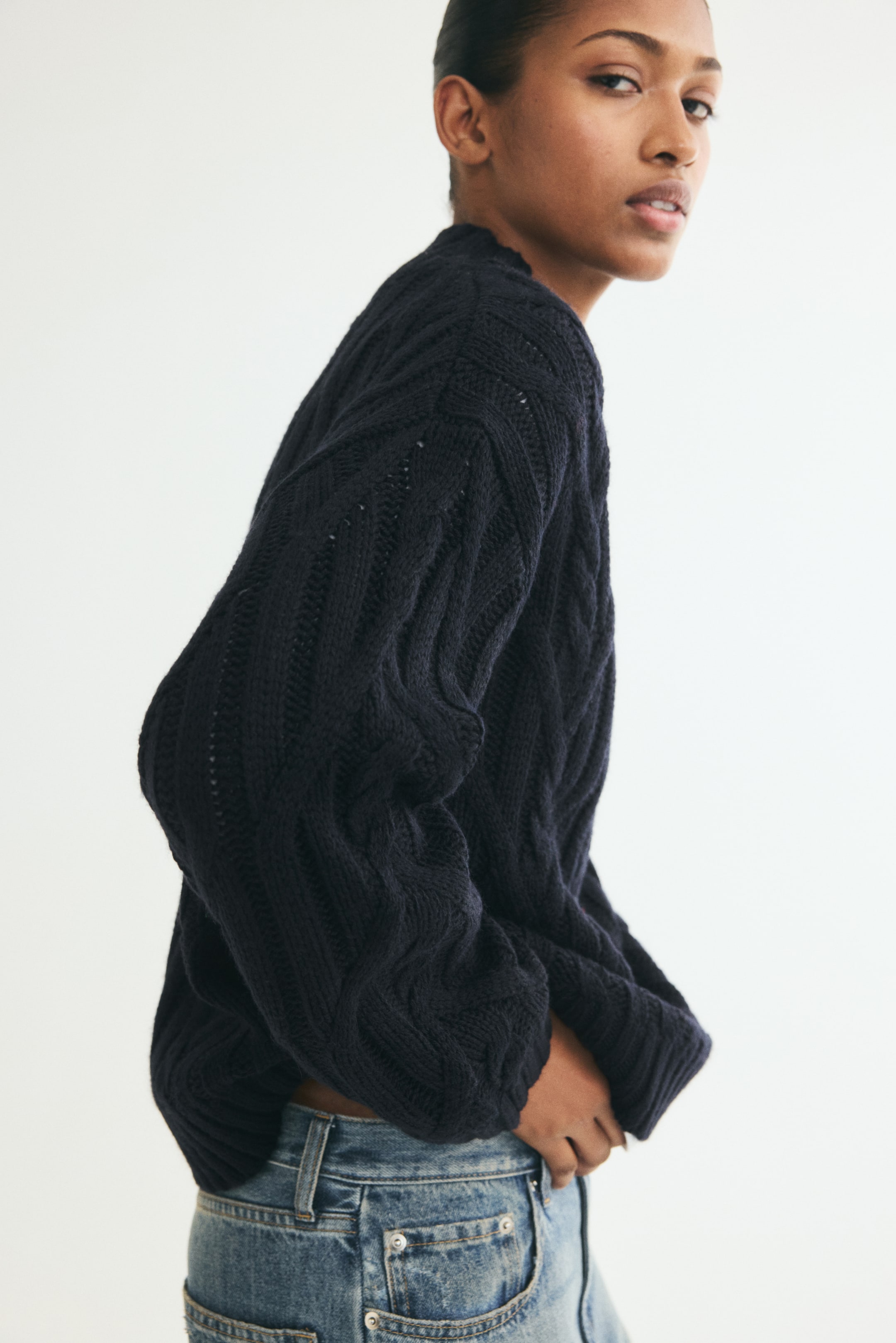 Ampliar la imagen: Se muestra un suéter tejido con trenzas en color azul marino oscuro y de corte oversized, visto de perfil lateral y orientado hacia la izquierda, resaltando su ajuste relajado con hombros caídos y mangas largas. El suéter presenta un patrón grueso de trenzas y detalles acanalados en el cuello, los puños y el dobladillo, que queda por encima de unos jeans azules de lavado claro.