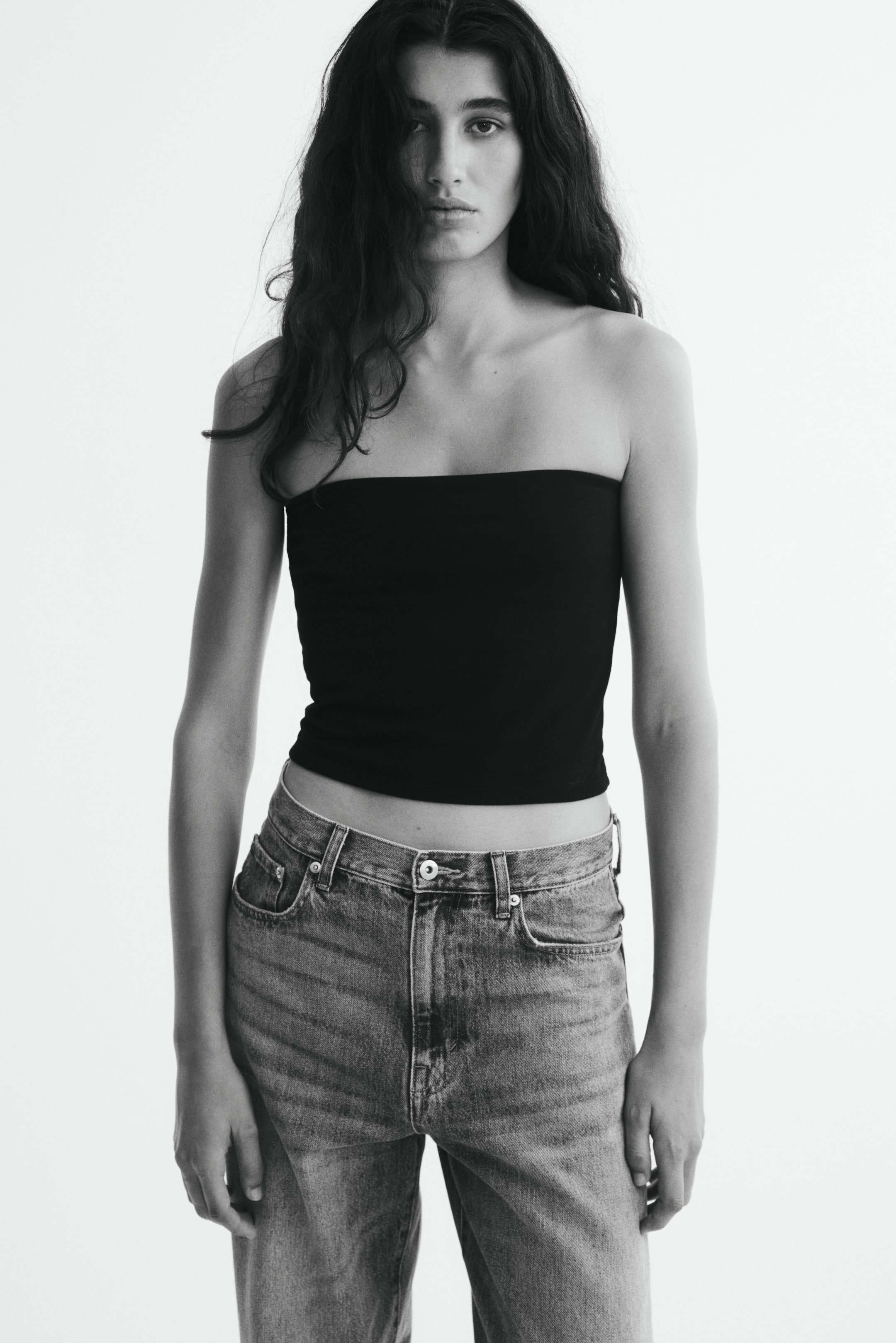 View larger image: The model is wearing a black, fitted strapless bandeau top paired with high-waisted denim jeans. The top features a straight-cut hem and fits snugly across the chest.