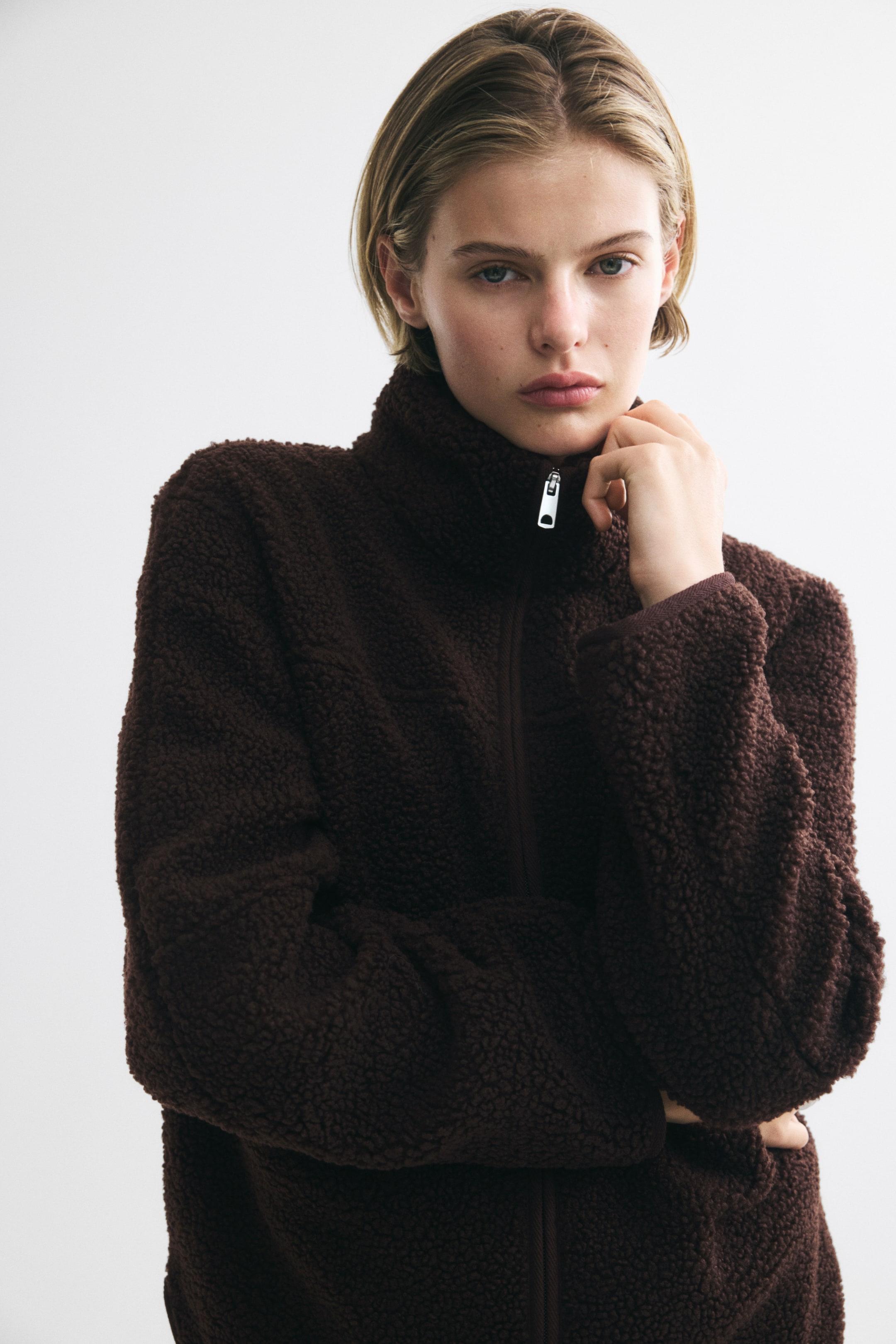 View larger image: A dark brown teddy fleece jacket with a stand collar and front zipper is being worn by the model. The model is posing with one hand resting near the collar and the other arm crossed over the torso, showcasing the jacket’s soft, textured fabric.