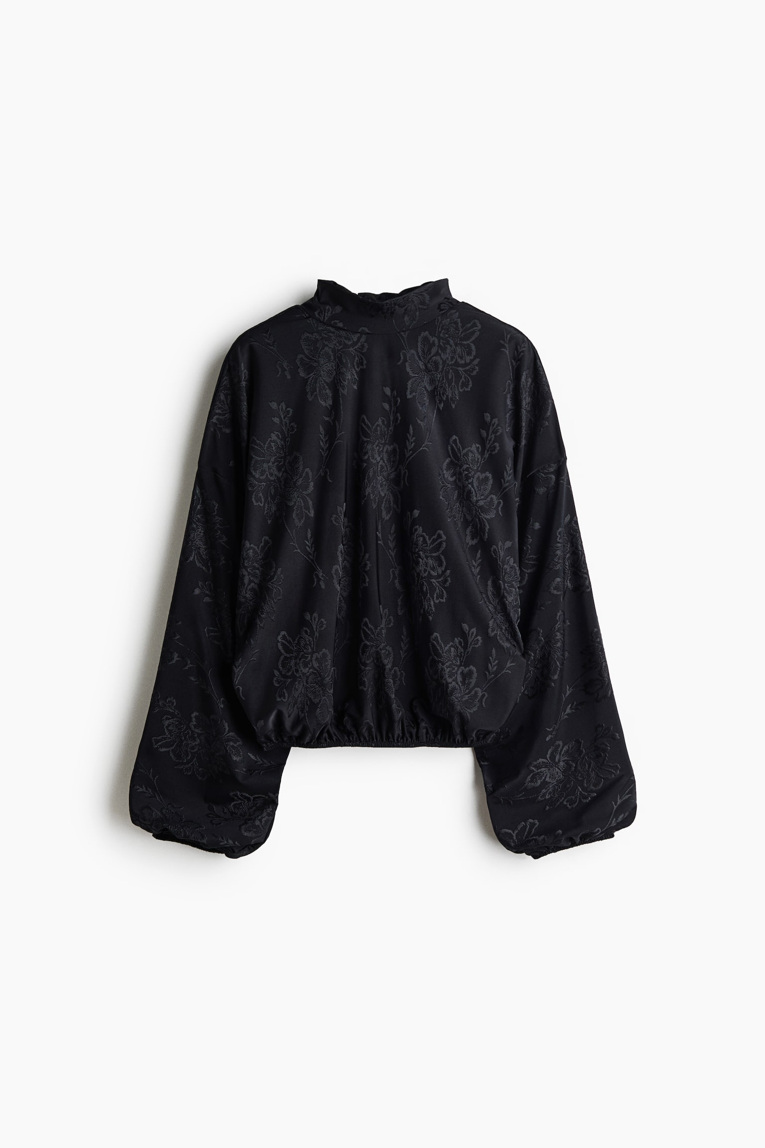 En svart, oversized blus i ett ton-i-ton-blommigt jacquardtyg visas framifrÃ¥n, med en hÃ¶g stÃ¥krage och voluminÃ¶sa ballongÃ¤rmar med resÃ¥r i Ã¤rmsluten. Blusens nederkant Ã¤r rynkad till en elastisk bubbelkant, vilket skapar en puffig silhuett.