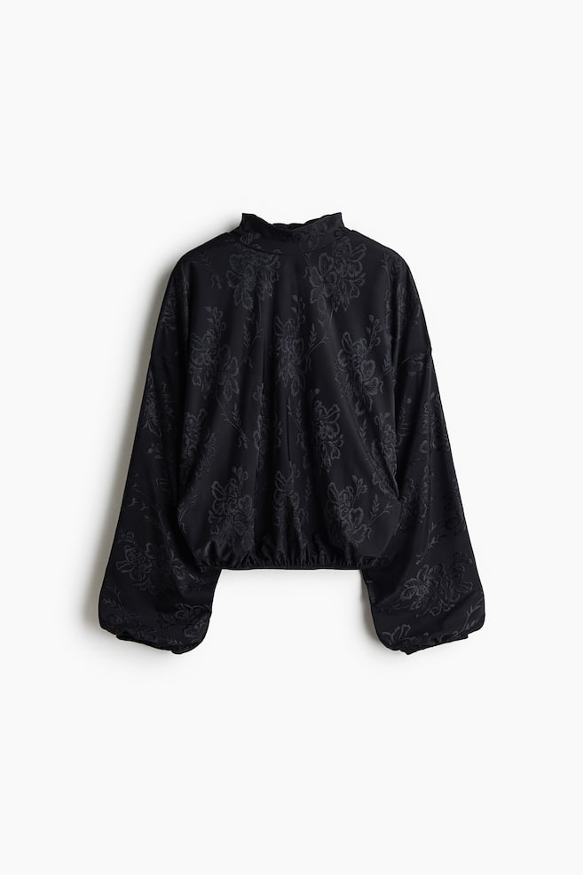 Eine schwarze, oversized Bluse aus tonalem Blumen-Jacquardstoff ist von vorne zu sehen und verfügt über einen hohen Stehkragen sowie voluminöse Ballonärmel mit elastischen Bündchen. Der Saum der Bluse ist zu einem elastischen Bubble-Saum gerafft und sorgt für eine voluminöse Silhouette. - 1