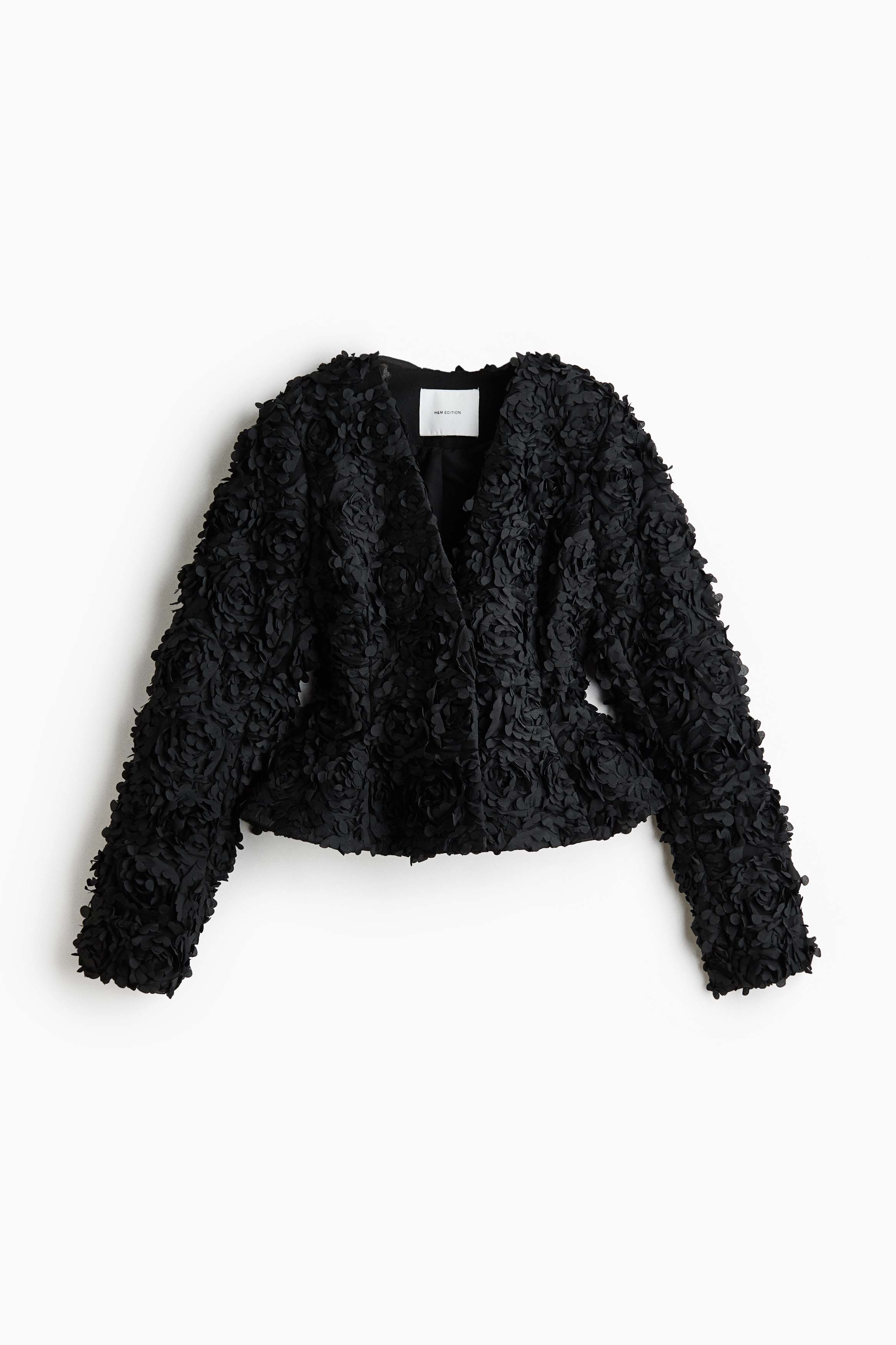 Une veste noire ornÃ©e dâappliquÃ©s floraux en relief, denses et tridimensionnels, ainsi que dâun col volantÃ© est portÃ©e par le mannequin. La veste est superposÃ©e Ã  une chemise noire Ã  volants et associÃ©e Ã  un pantalon noir.
