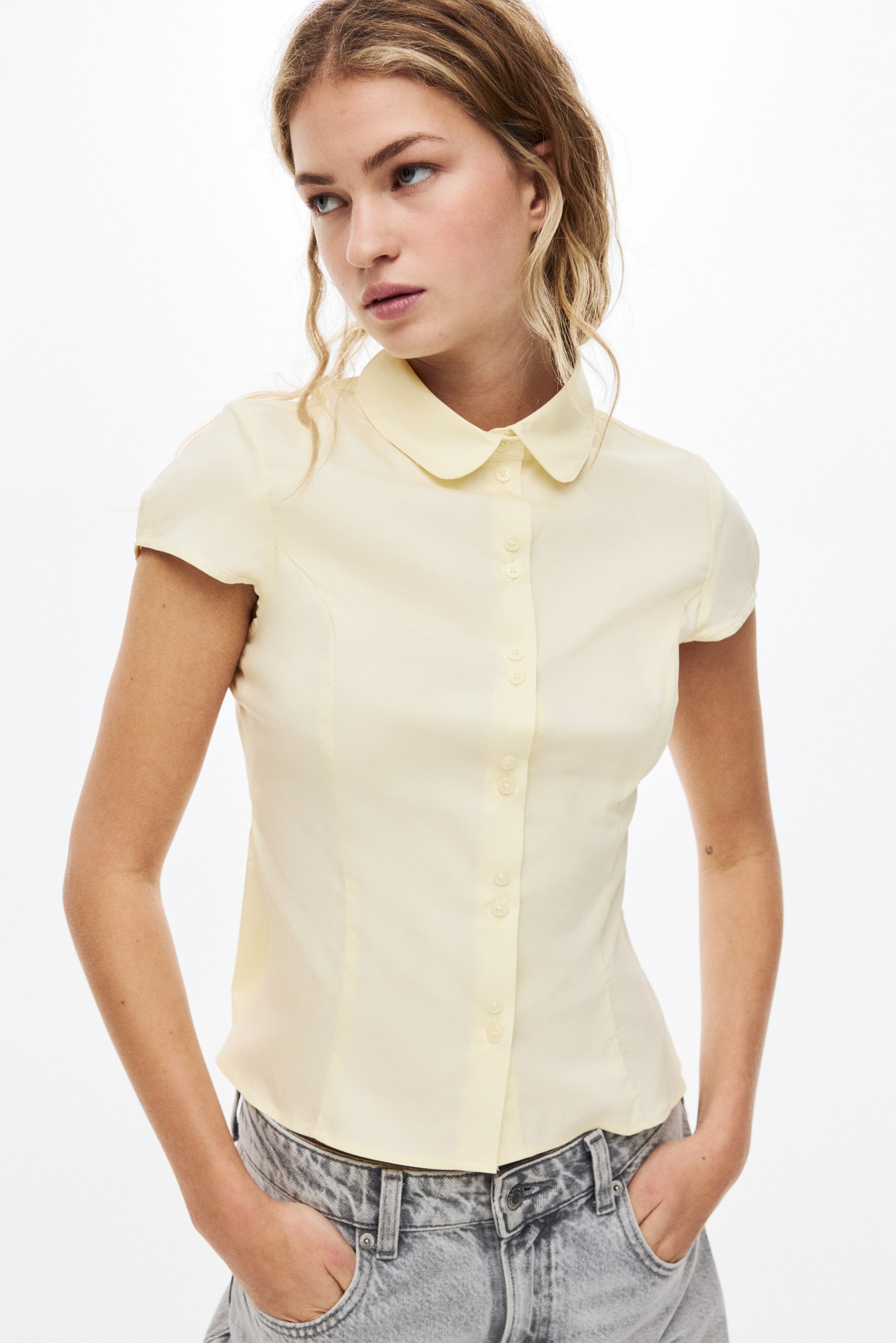 View larger image: A light yellow short-sleeved shirt featuring a rounded Peter Pan collar and a full button-front closure with matching buttons. It showcases a fitted silhouette with shaping seams, and its short sleeves end mid-bicep.