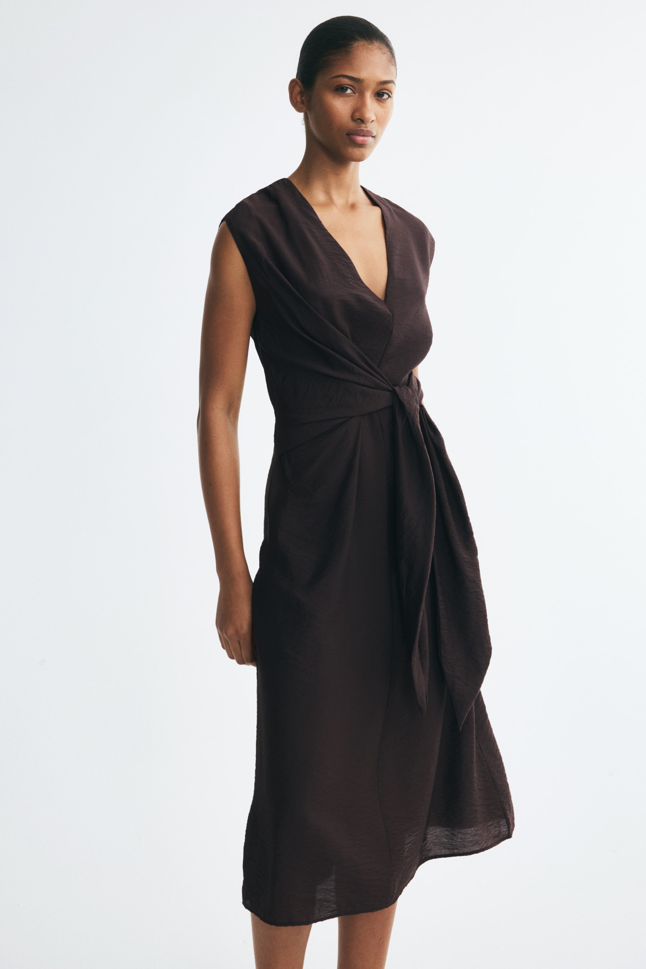 View larger image: The model is wearing a dark brown satin dress with a sleeveless design and a V-neckline. The dress features a draped bodice gathered into a twisted knot detail at the waist, with long fabric ties hanging down the front.