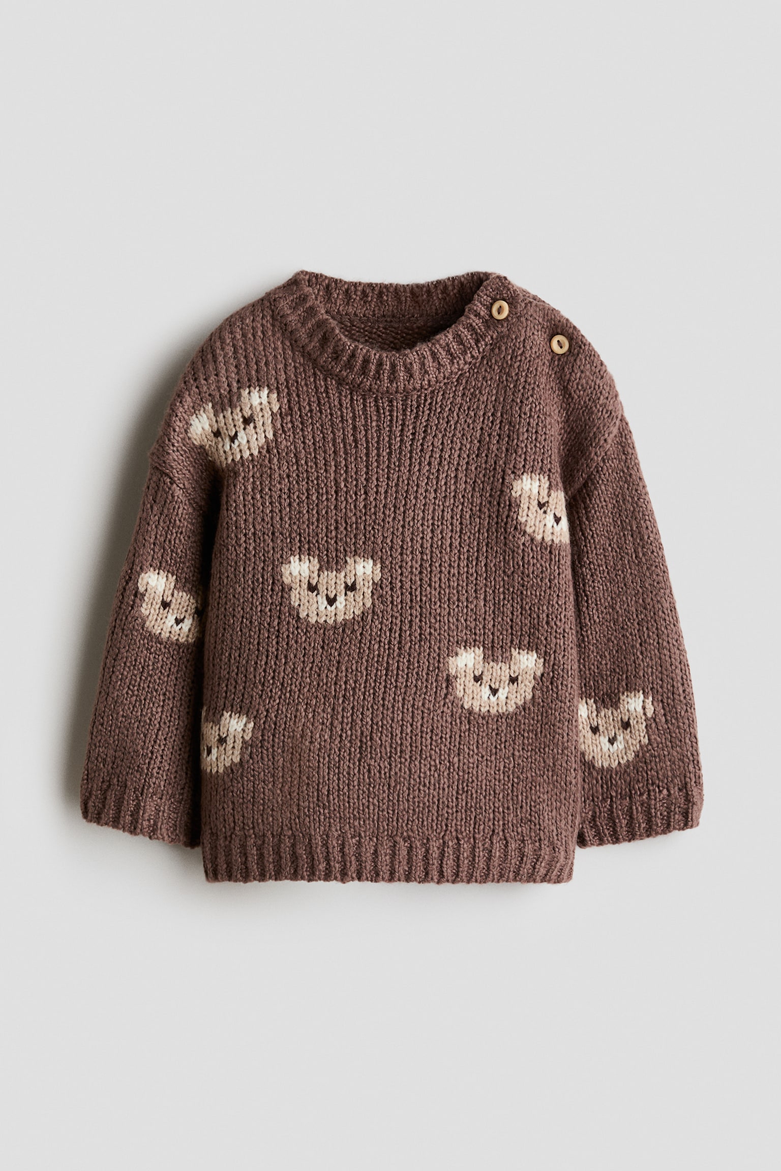 A brown jacquard-knit jumper is shown from the front, featuring a pattern of scattered beige knitted bear faces. It has a round, rib-trimmed neckline with two button fasteners on one shoulder, alongside wide ribbed trims at the cuffs and hem.