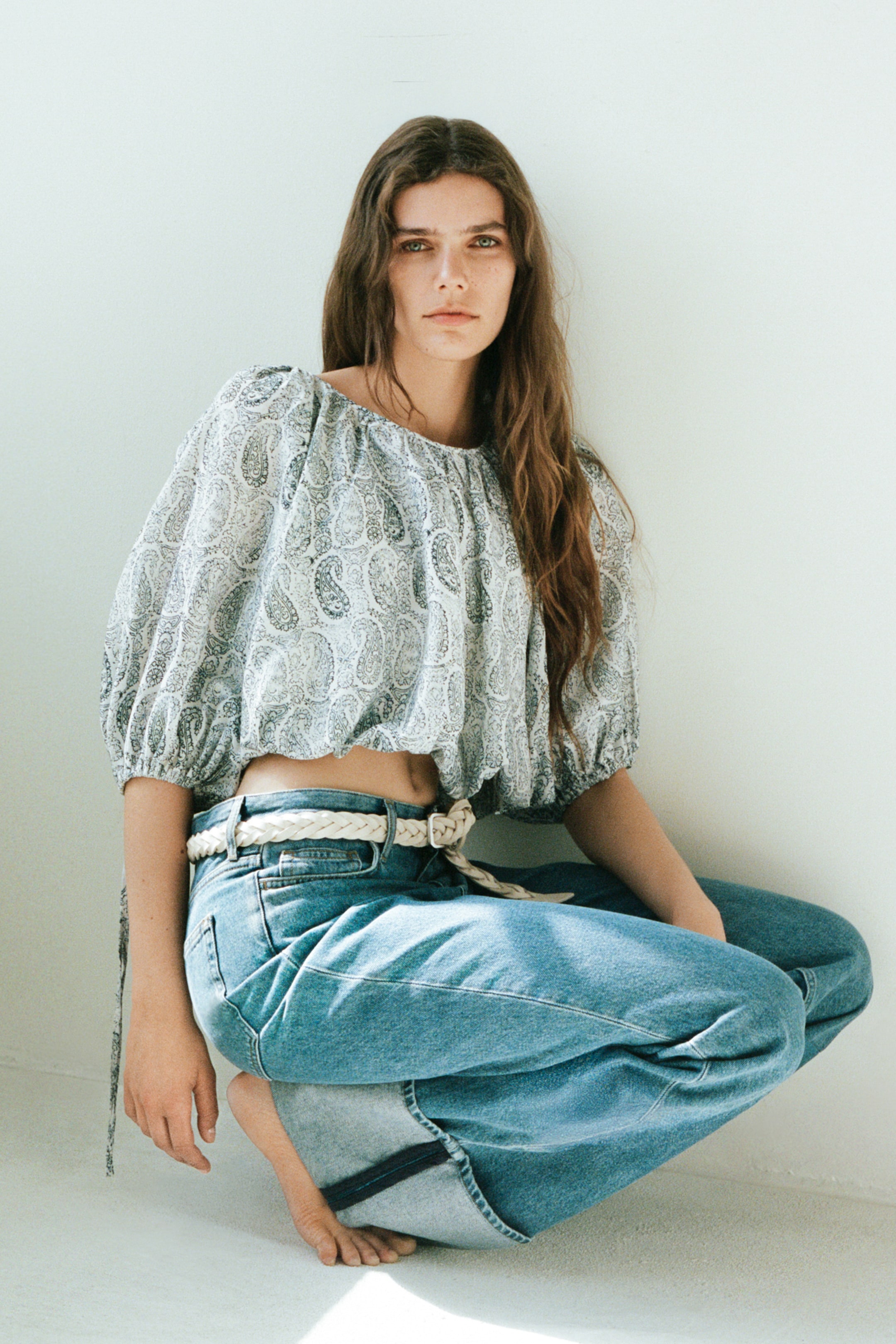 Xem ảnh lớn hơn: Một chiếc áo crop top với họa tiết paisley nhẹ nhàng, cổ áo co giãn, và tay áo phồng dài đến khuỷu tay đang được kết hợp với quần jeans xanh cạp cao, ống rộng. Một chiếc thắt lưng bện nhẹ đang thắt chặt quần jeans ở eo.