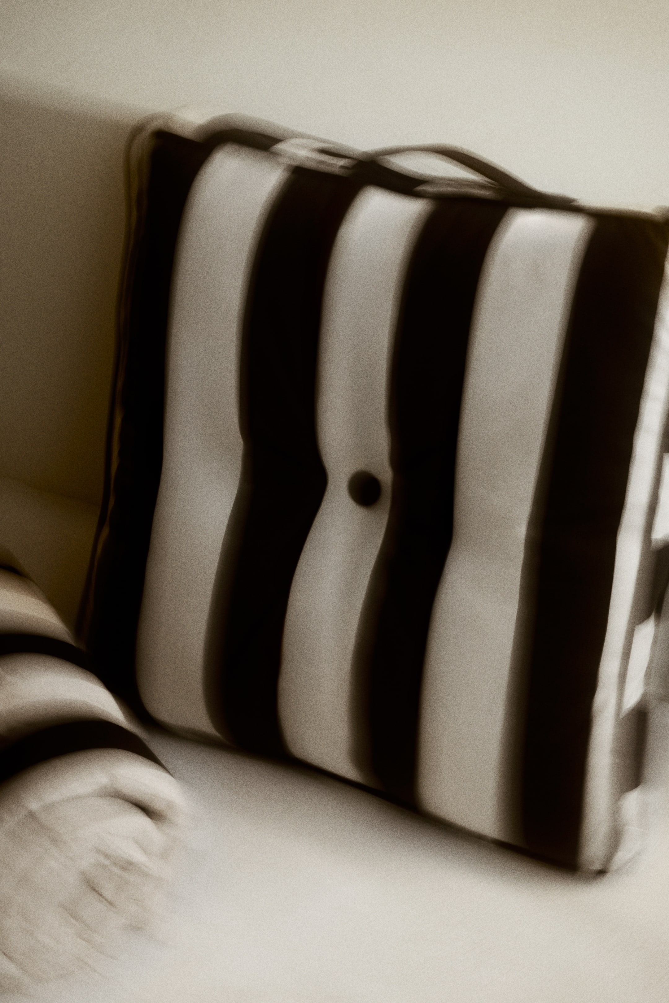 Agrandir l'image: Un coussin présente des rayures verticales à fort contraste dans des tons sombres et clairs, et arbore un design capitonné avec un bouton sombre au centre. Une poignée de transport est positionnée le long de son bord supérieur, et le coussin est légèrement orienté vers la droite.