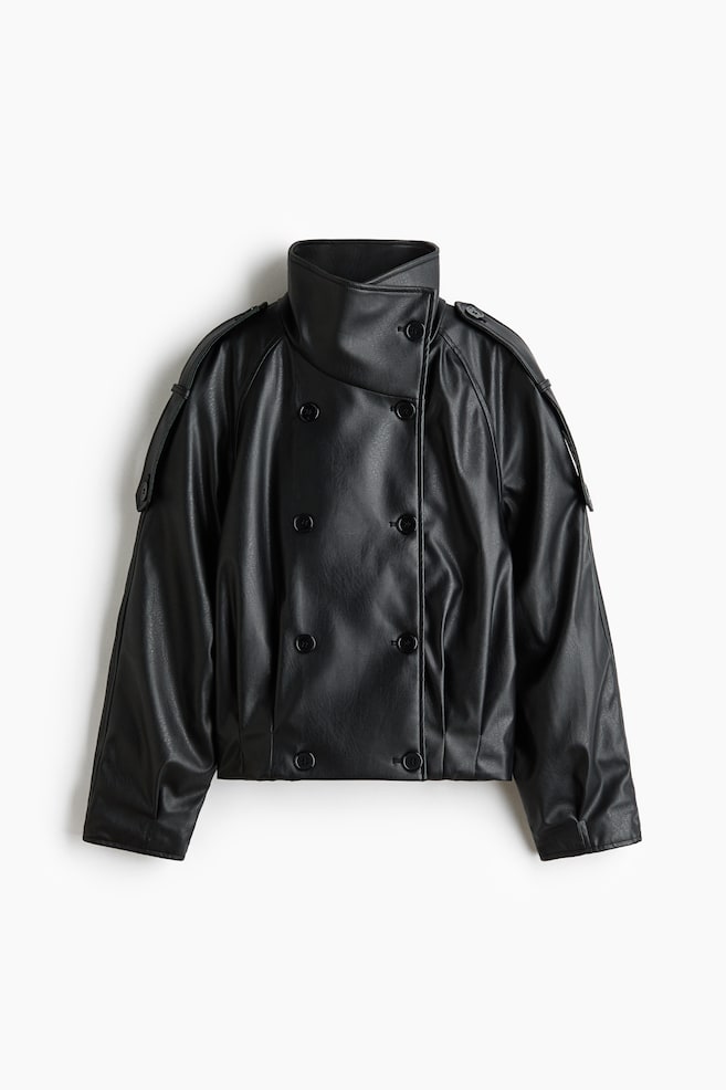 An oversized, double-breasted black jacket in a smooth, leather-like fabric is shown from a front-facing perspective, featuring a high funnel collar with a button fastener. The jacket features raglan sleeves with buttoned shoulder tabs extending down the upper arms, and pleating at the hem that creates a slightly puffed silhouette. - 1