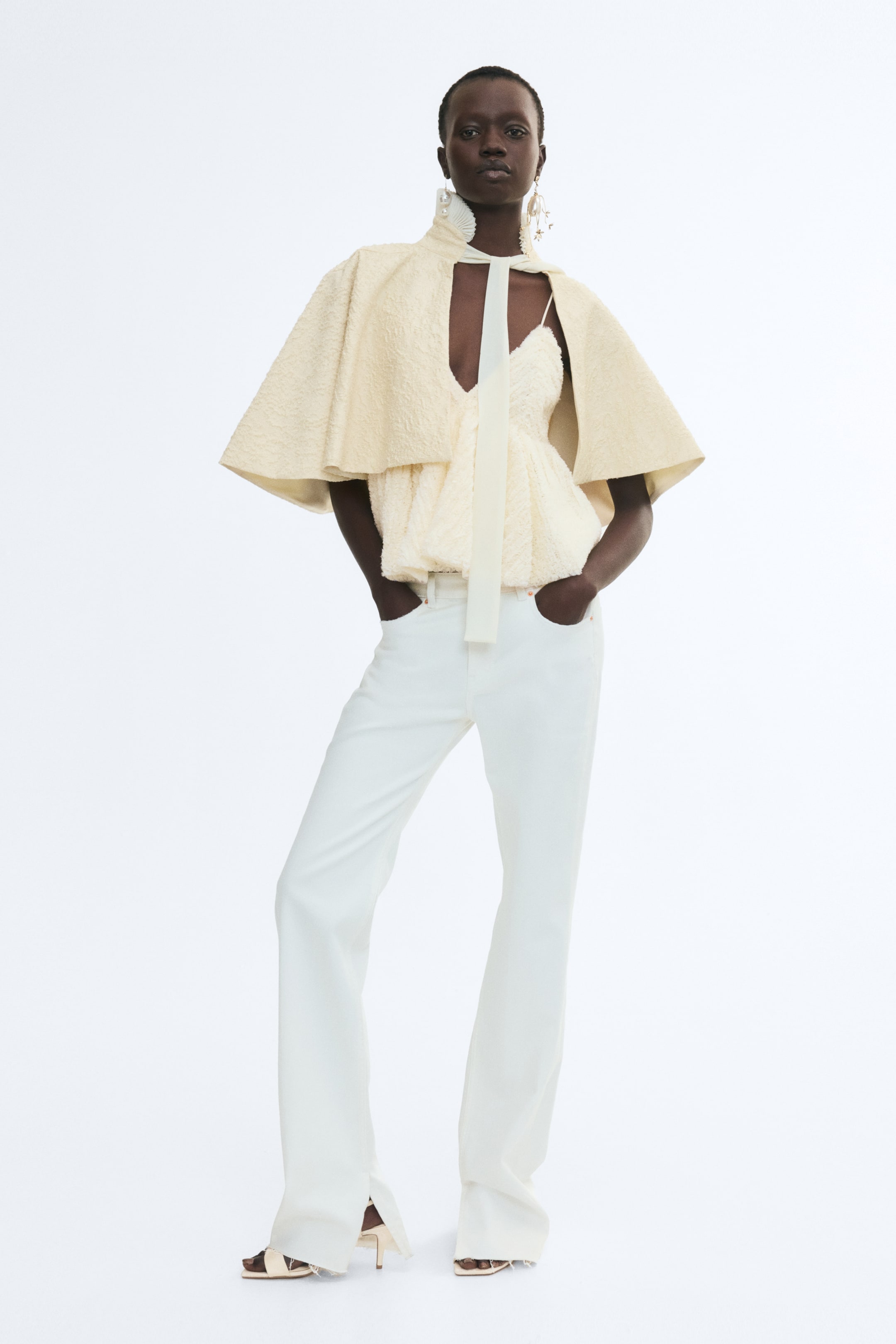 Visa större bild: En krämfärgad volangprydd camisole med V-ringning och peplum-silhuett bärs av modellen. Camisolen bärs under en matchande strukturerad capelet och matchas med vida vita byxor.