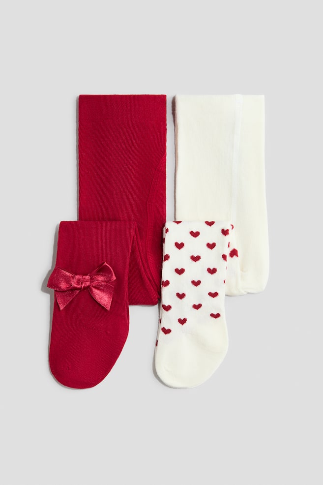 Two pairs of fine-knit cotton-blend tights are shown folded in a front-facing view. The pair on the left features a solid red colour with a matching satin bow detail near the foot, while the pair on the right features an off-white colour with one leg covered in an all-over pattern of small red hearts. - 1