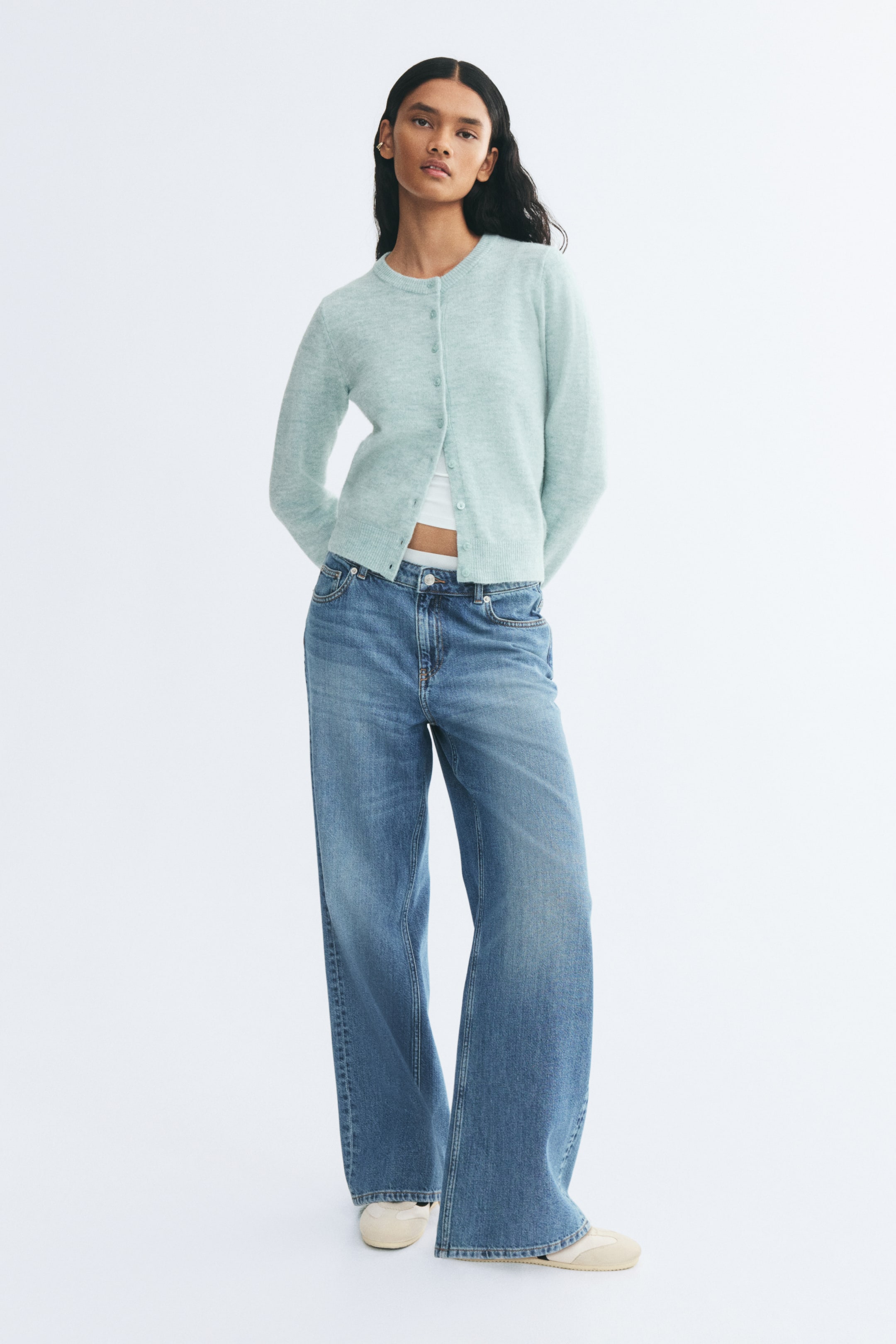 View larger image: A light green, fitted knit cardigan with a round rib-trimmed neckline, ribbed cuffs and hem, and a button-front closure is worn by the model. The cardigan is styled partially unbuttoned at the hem over a white top and paired with wide-leg blue jeans.