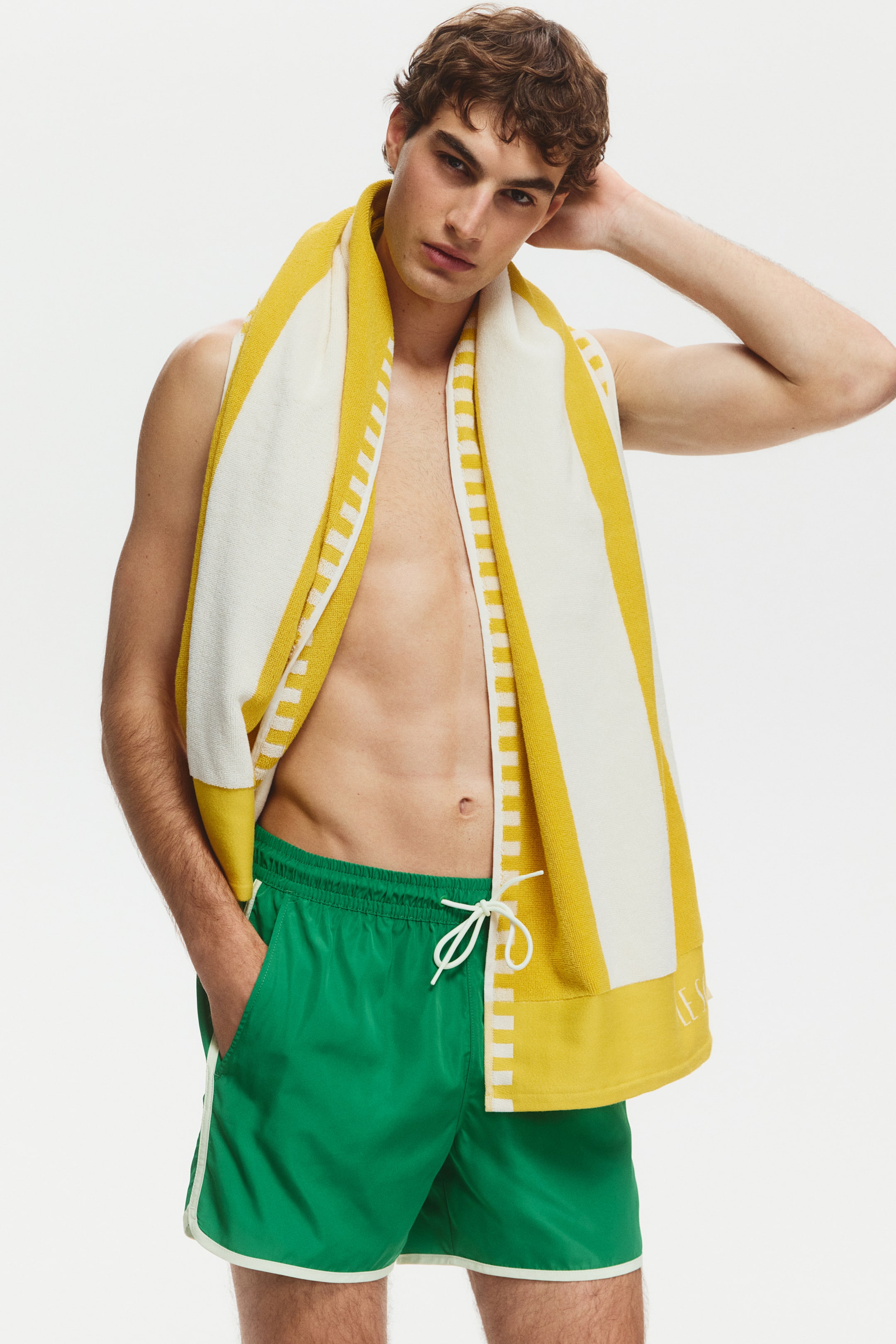 View larger image: The model is wearing green swim shorts. These shorts feature an elastic waistband with a white drawstring and white piping along the side seams and hem. The model's hand rests in a side pocket.