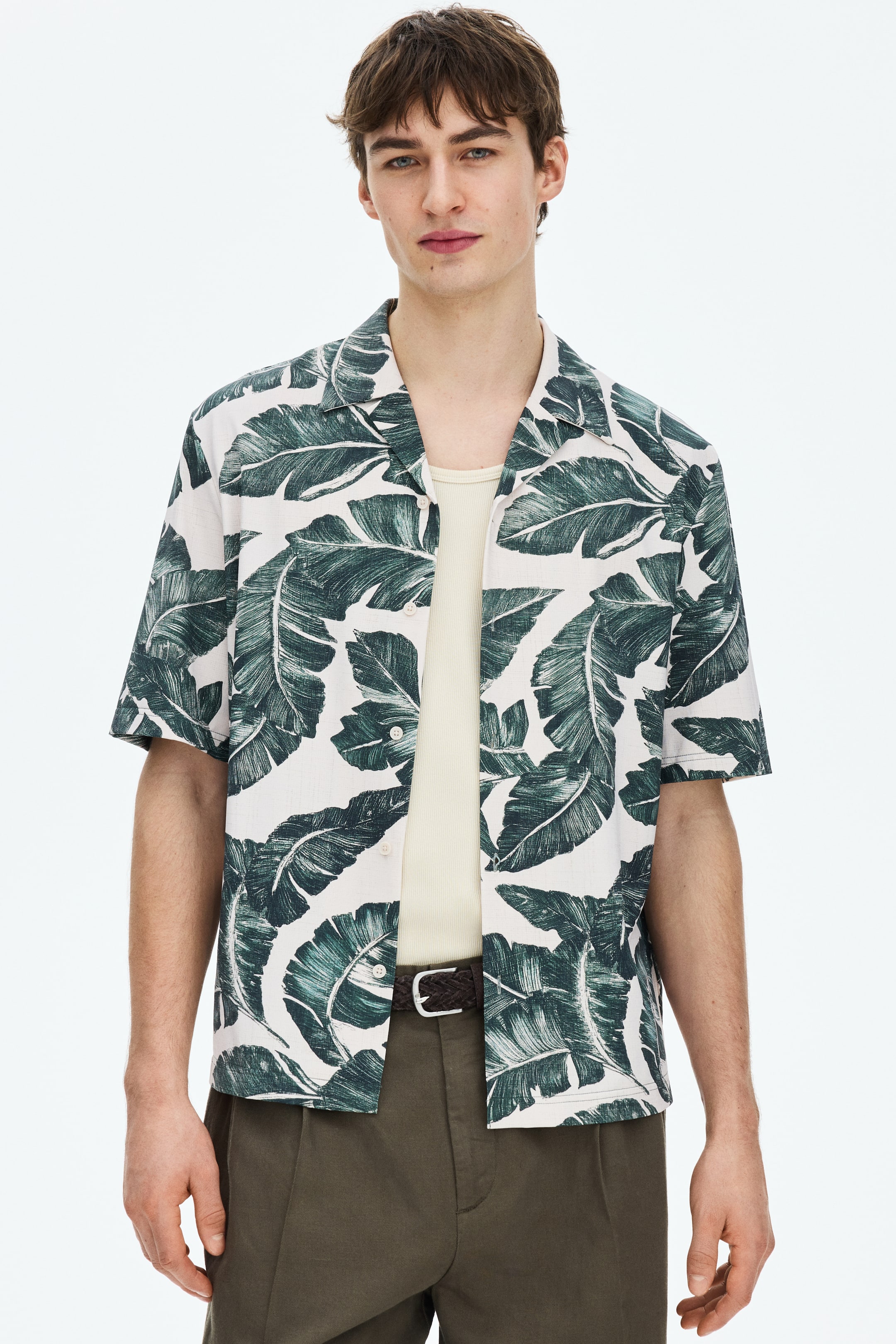View larger image: A short-sleeved shirt showcasing a dark green tropical leaf print on a white background. It features a camp collar and is worn unbuttoned over a light-coloured undershirt.