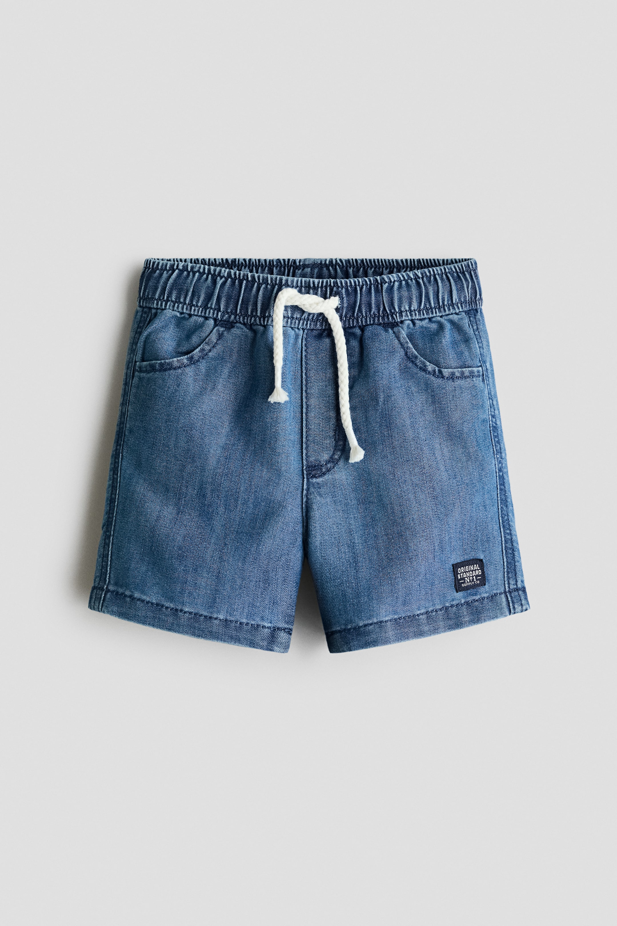 View larger image: Blue denim shorts are displayed from the front, featuring an elasticated waistband with a white drawstring knotted at the centre. They are designed with two rounded front pockets and a stitched hem, and a small dark label with white text is visible on the lower right leg.