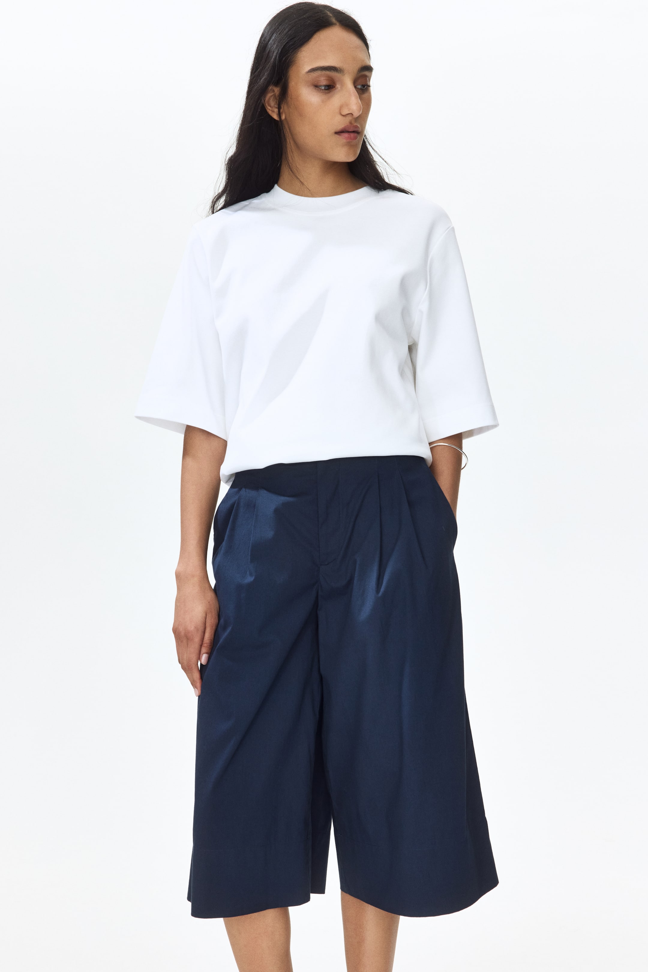 View larger image: A white short-sleeved t-shirt showcases a crew neck and broad sleeves, neatly tucked into navy blue culottes. The culottes are crafted with a high-waist, front pleats, and wide, cropped legs that end at mid-calf, also featuring side pockets.