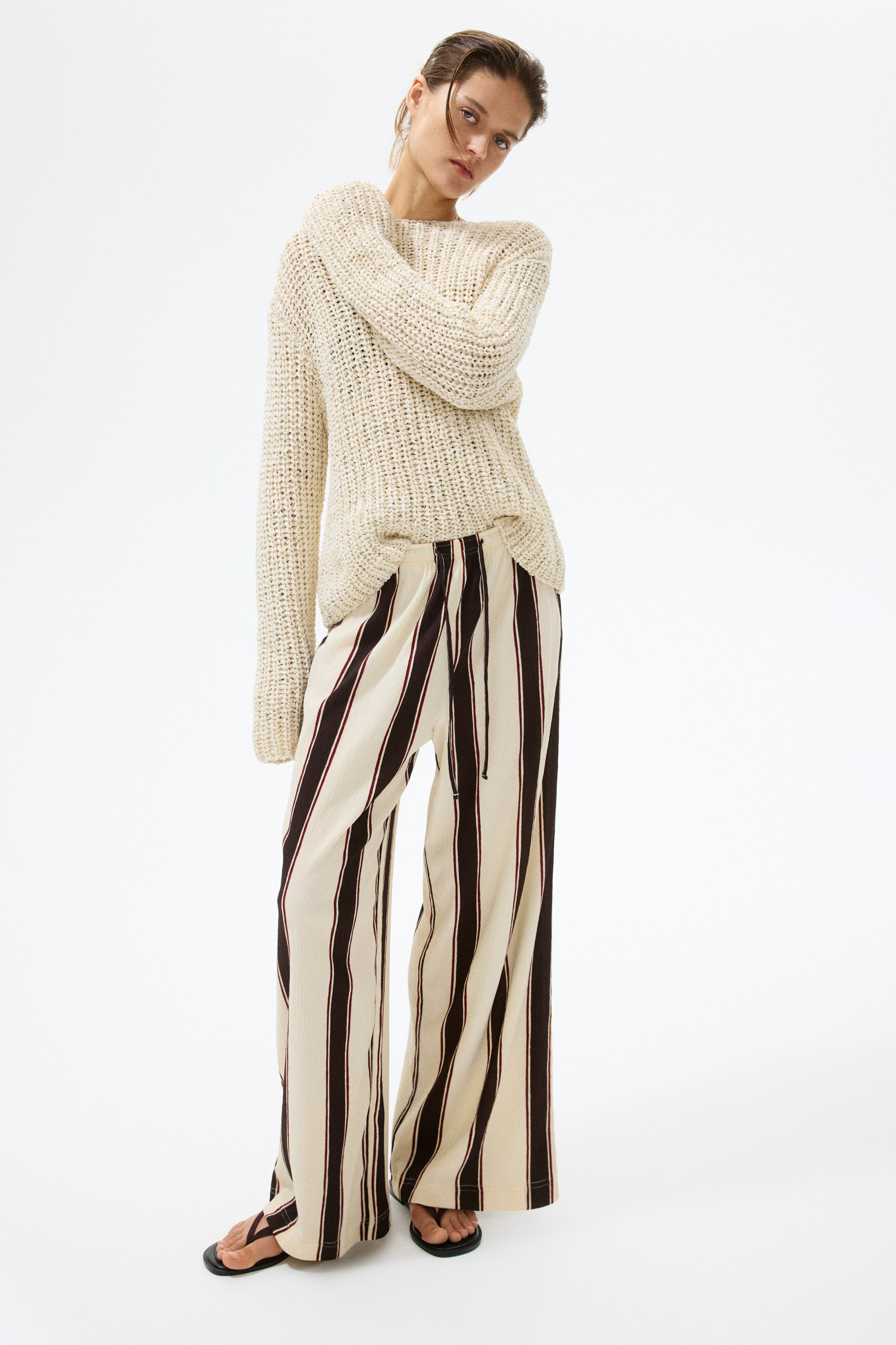 Ampliar la imagen: Un suéter de punto grueso color crema se lleva sobre pantalones de pierna ancha con rayas verticales en crema y marrón oscuro. El suéter de corte holgado presenta un cuello redondo y mangas largas, mientras que los pantalones exhiben una cintura con cordón y una silueta fluida, combinados con sandalias de tanga negras.