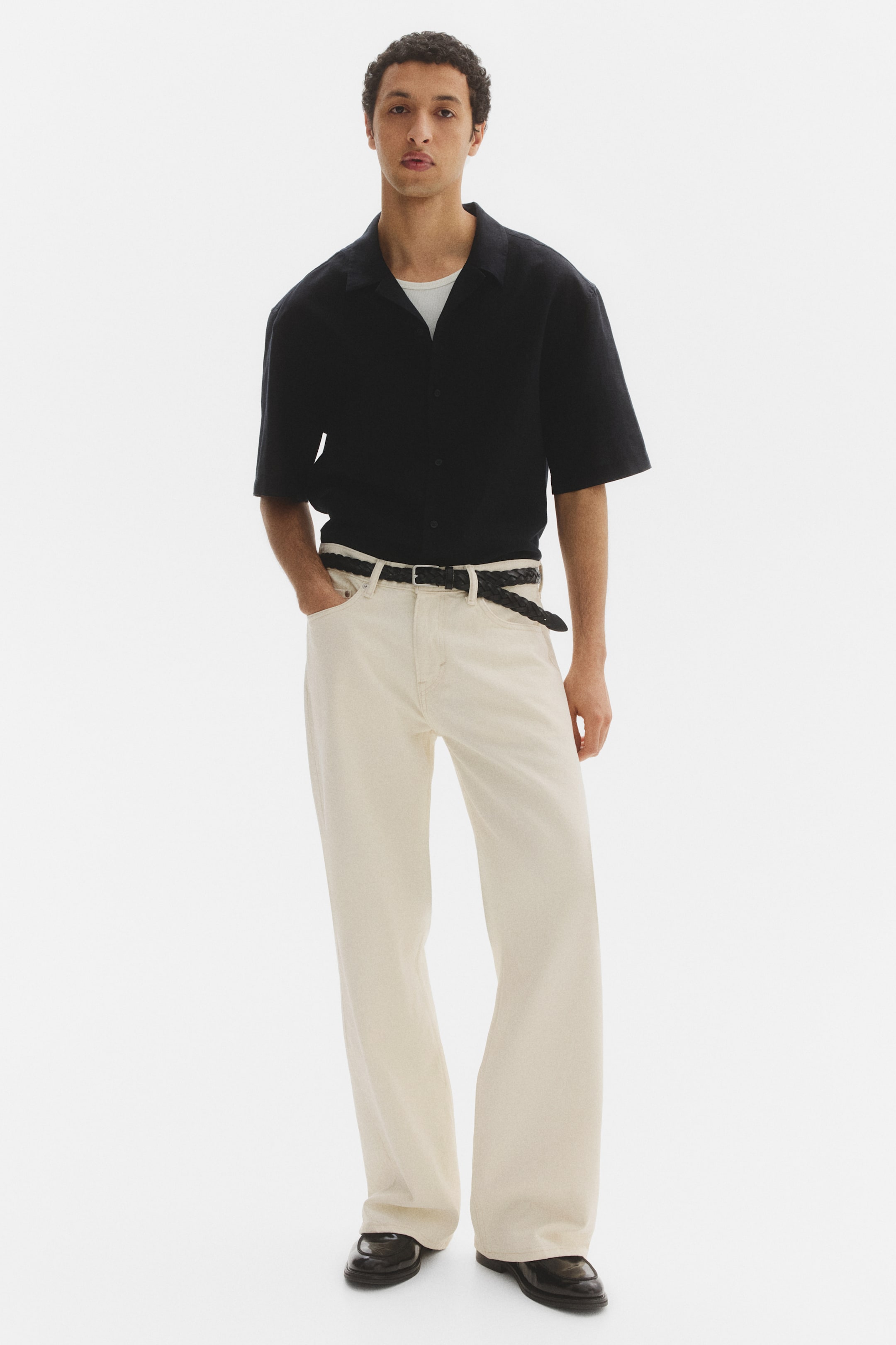 View larger image: A black short-sleeved camp collar shirt is styled open over a white crew neck t-shirt, teamed with light cream wide-leg trousers secured by a black braided belt, and black loafers complete the look.
