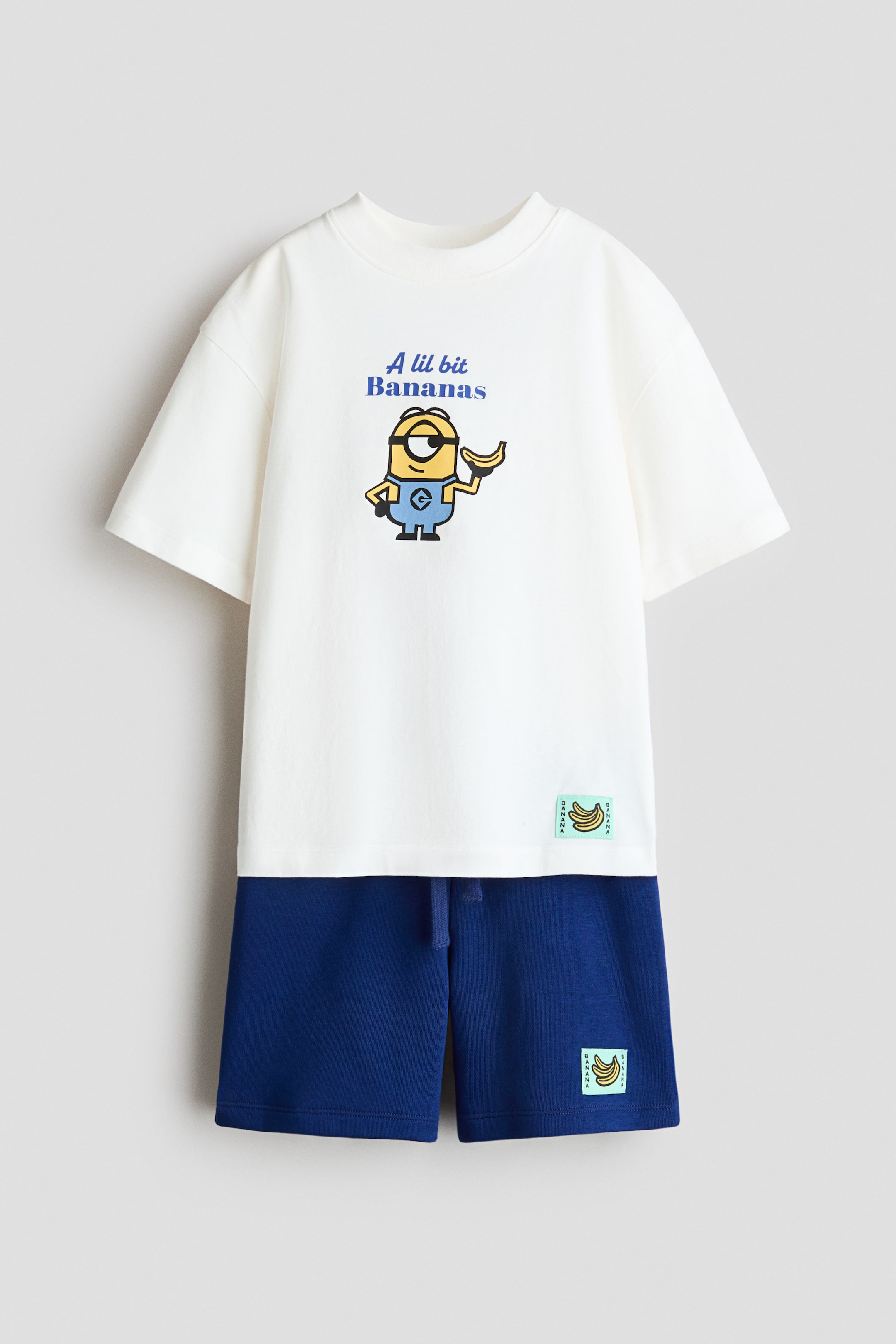 Agrandir l'image: L'ensemble assorti présente un T-shirt blanc cassé et un short bleu foncé, tous deux montrés de face. Le T-shirt arbore un personnage Minion tenant une banane et le texte "A lil bit Bananas", avec des épaules tombantes, des manches larges et un col côtelé. Le short affiche une taille à cordon de serrage, et les deux pièces sont agrémentées d'une petite étiquette sur le thème de la banane.