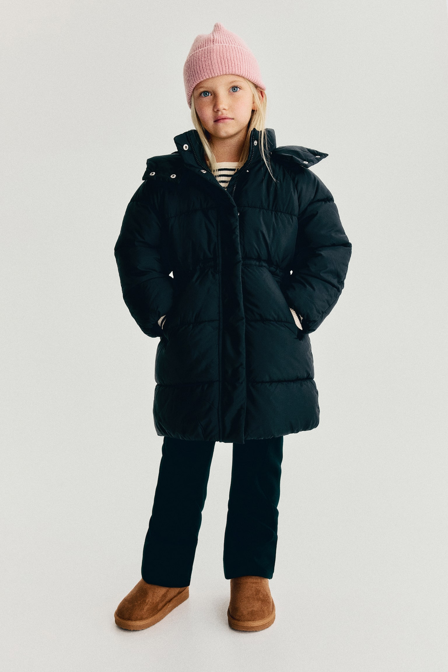 A black mid-length puffer jacket is shown facing front, featuring horizontal quilting, an attached hood with a high collar, and a concealed front fastening. The jacket has a gathered, elasticated waist and two angled welt pockets on the lower front. - 1
