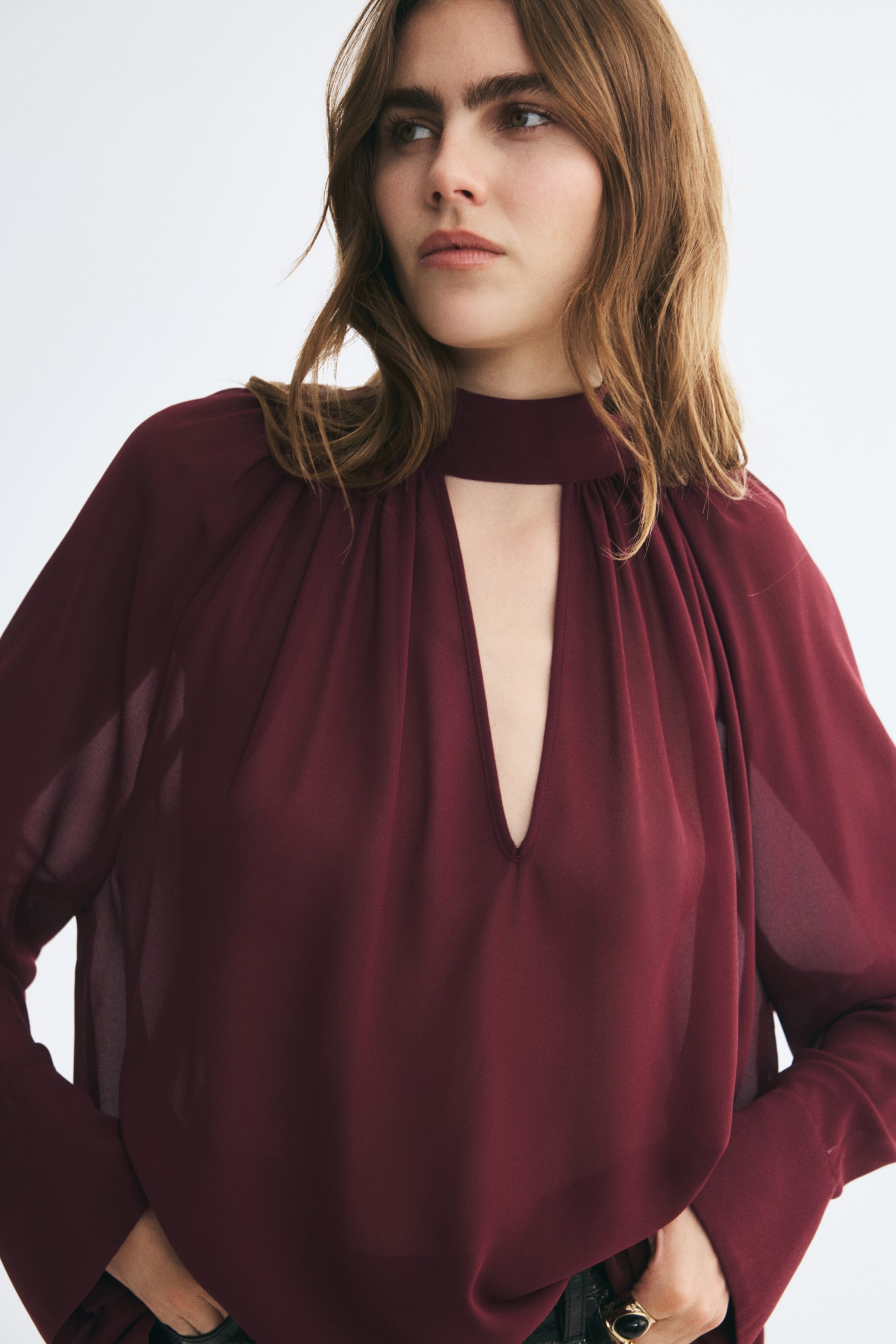 Voir une image plus grande: Une blouse bourgogne surdimensionnée en tissu transparent est portée par le mannequin, avec un col montant façon foulard, des fronces et une profonde ouverture en V à l’avant. La blouse est portée avec un pantalon foncé, dans lequel le mannequin glisse ses mains.