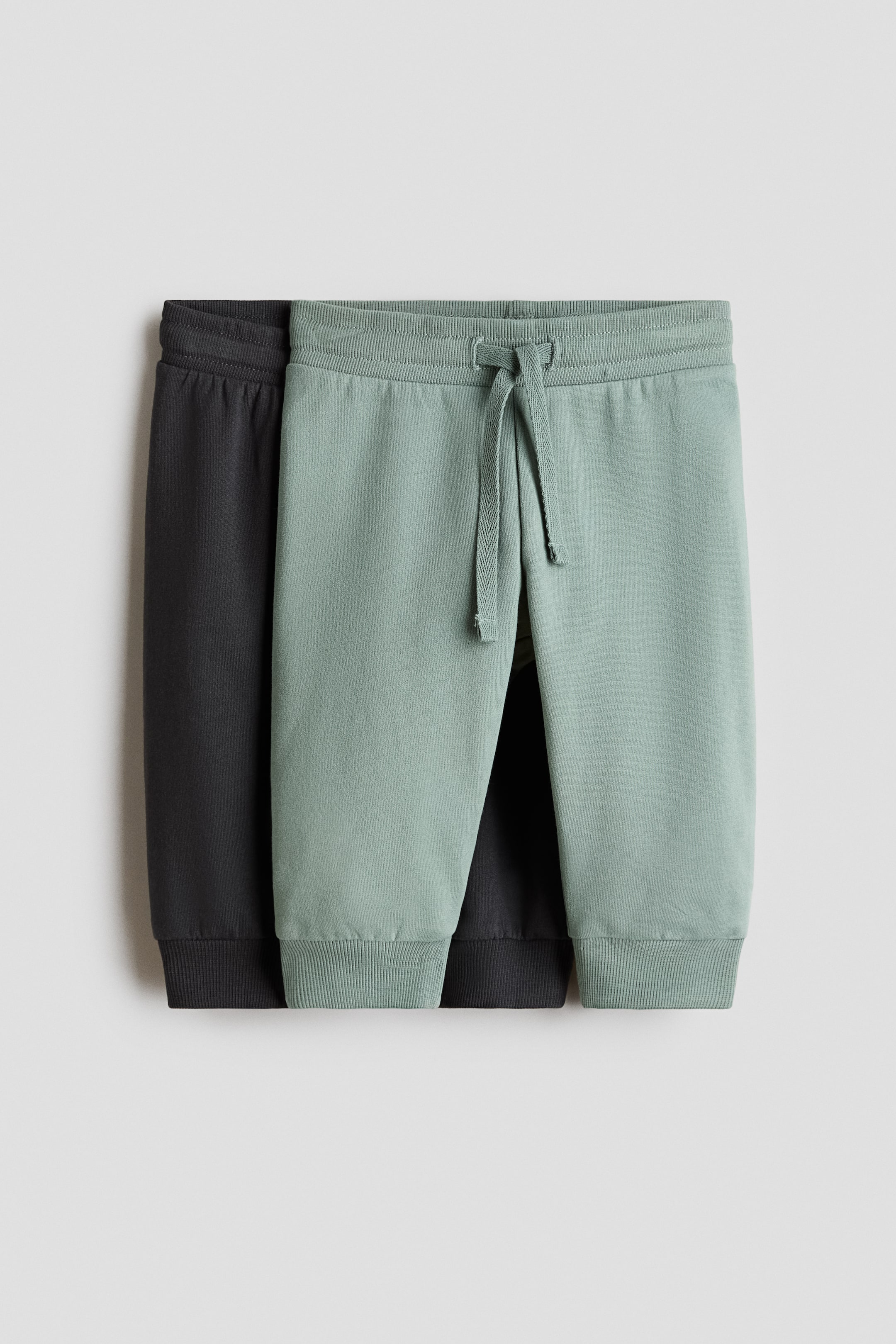 View larger image: Two pairs of sweatpants are displayed, with a light green pair partially overlapping a dark gray pair. Both pairs face forward, showcasing ribbed elastic waistbands and ribbed ankle cuffs. The light green pair also features a tied drawstring at the waist.