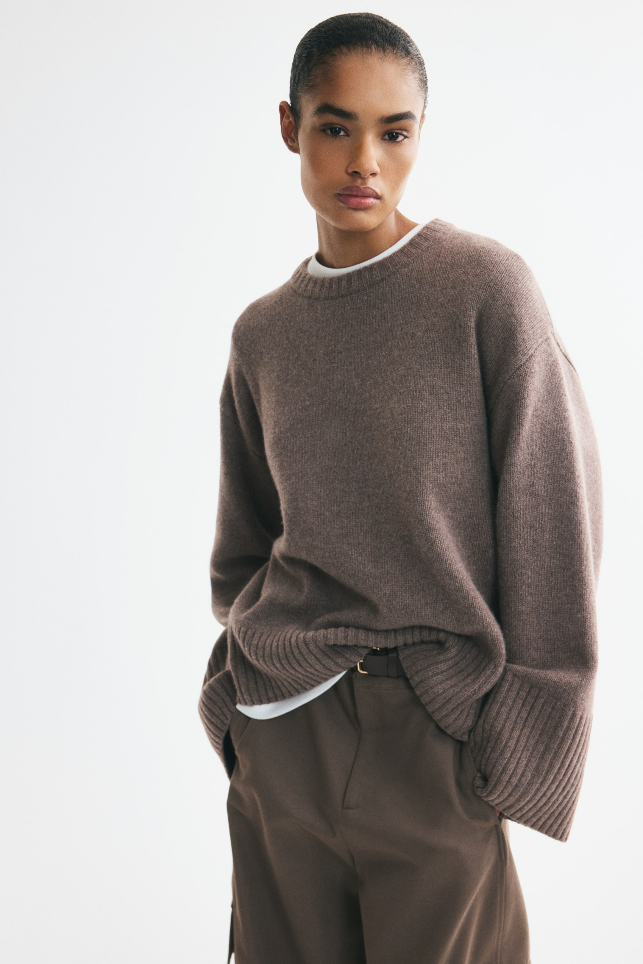 View larger image: The model is wearing an oversized brown jumper featuring dropped shoulders, extra-long sleeves, and ribbing at the neckline, cuffs, and hem. Underneath the jumper, a white t-shirt is layered.