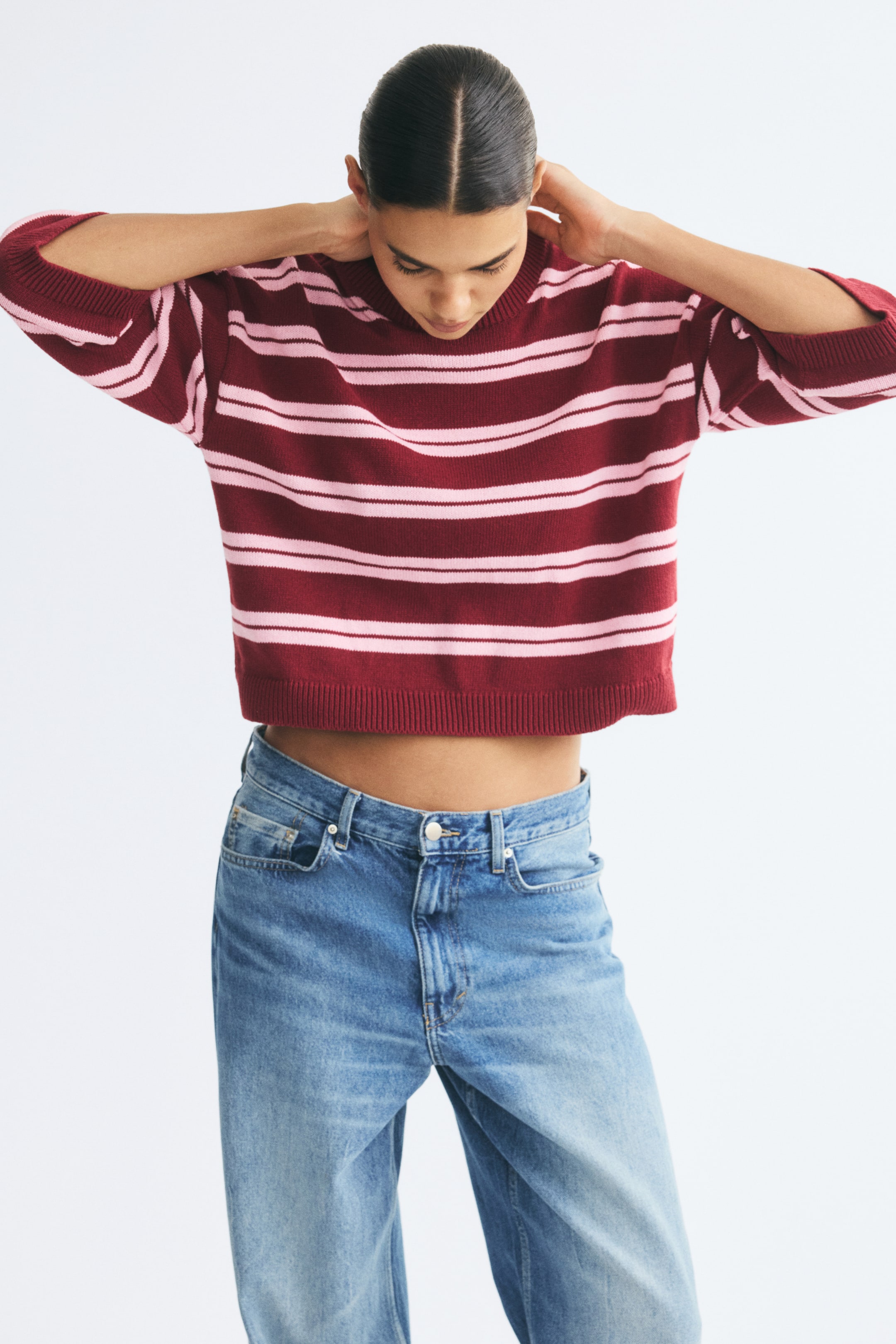 Visualizza immagine più grande: Una t-shirt corta in maglia con righe orizzontali bordeaux e rosa indossata dalla modella, abbinata a jeans blu. Il top crop presenta un girocollo rifinito a coste, spalle scese, maniche a 3/4 e bordi a coste su polsini e orlo.