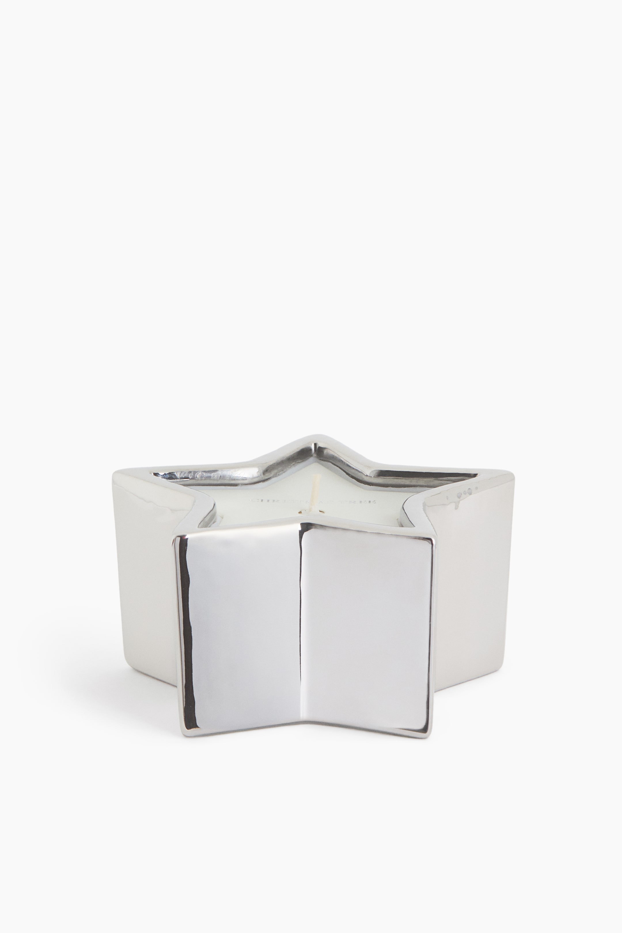 View larger image: A star-shaped candle is placed in a reflective metallic holder with a silver finish and a five-point star silhouette, shown from a front-facing angle. The polished container holds white wax with a single wick visible at the centre.