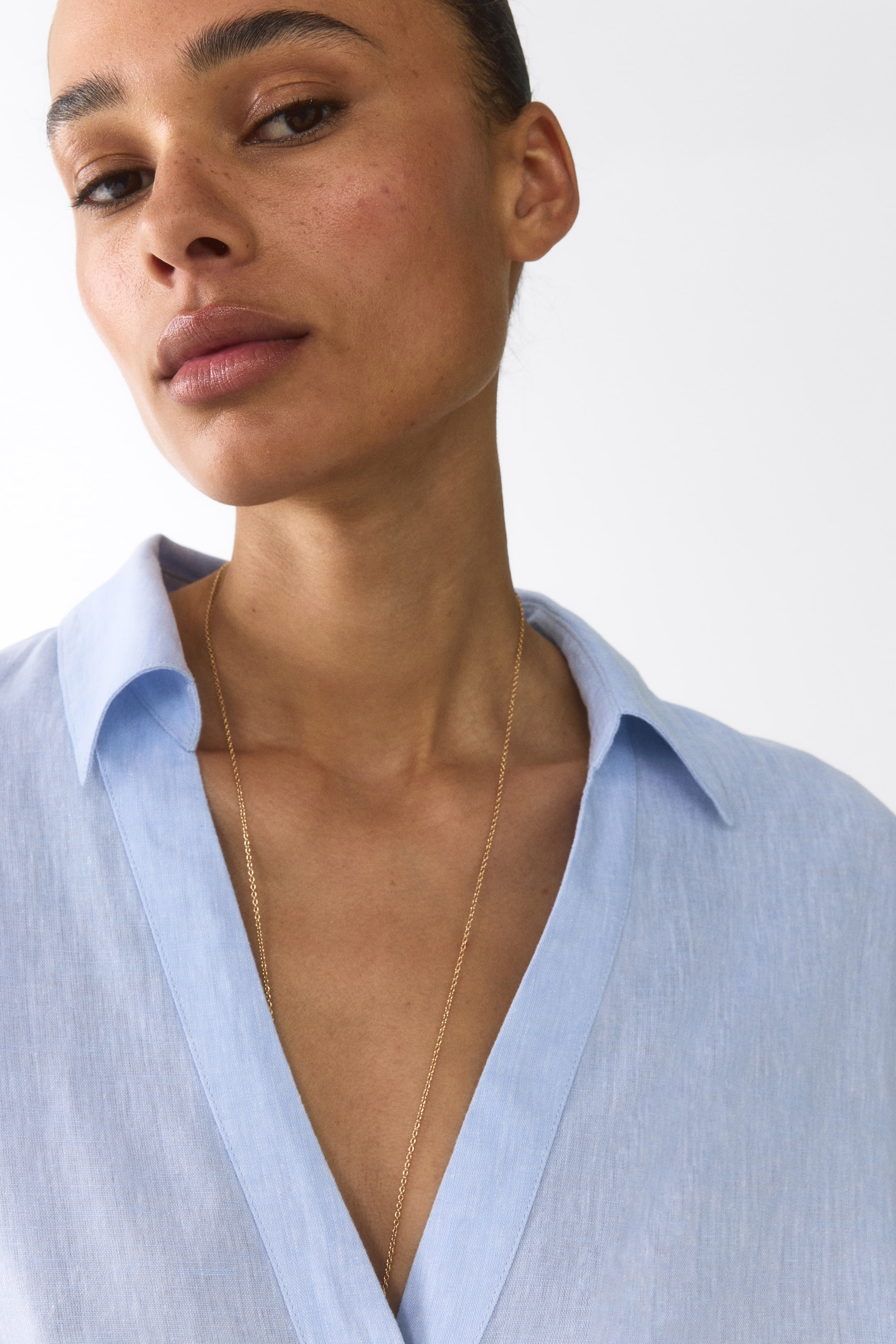 View larger image: A light blue linen shirt featuring a classic pointed collar and a deep V-neck opening. A thin gold chain necklace is hanging centrally within the V-neck.