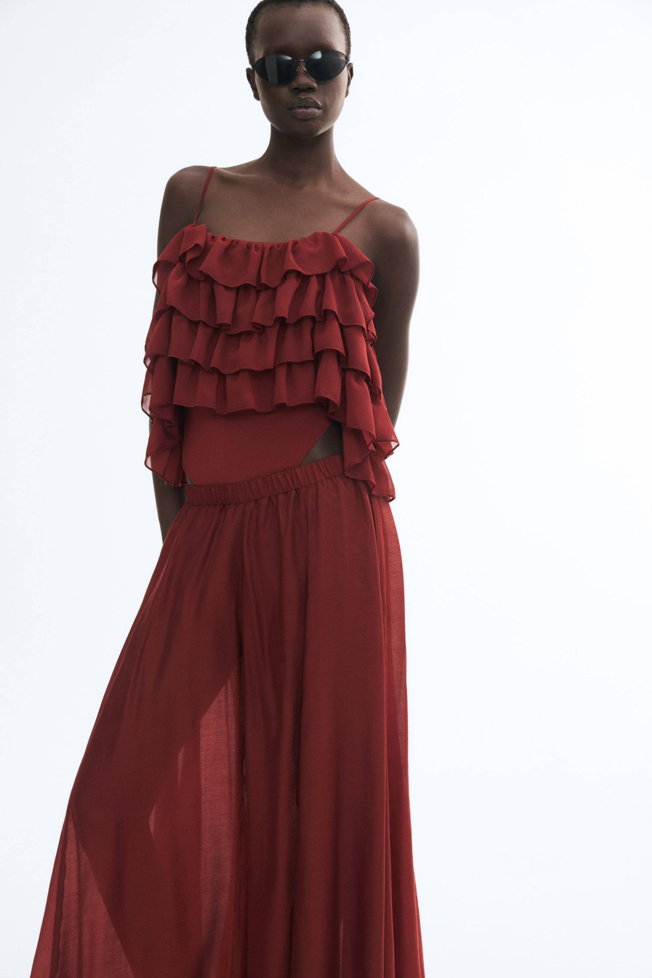 View larger image: A rust-red bodysuit featuring narrow shoulder straps and a tiered, ruffled mesh overlay is being worn by the model. The bodysuit is styled with matching sheer, wide-leg trousers.