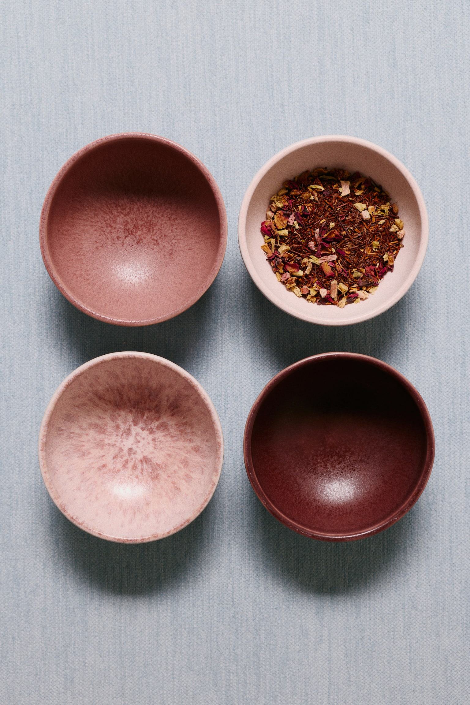 Four stoneware bowls display a reactive glaze in varying shades of pink, mauve, and deep brown, with three of the bowls stacked and one bowl positioned to the right. The bowls feature a classic rounded shape with a footed base and a textured, variegated surface finish. - 1