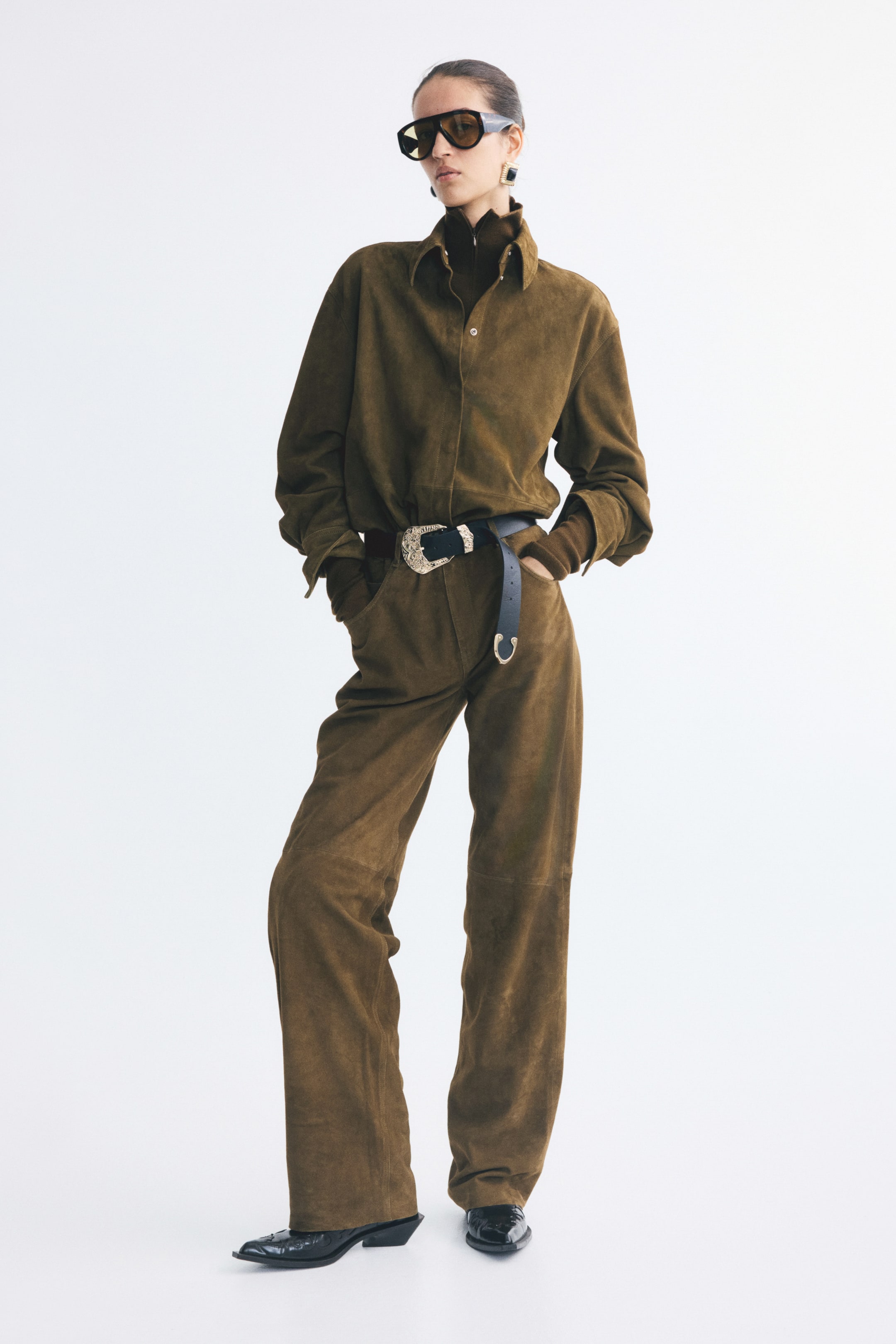 Agrandir l'image: Un pantalon cinq poches en daim marron, à coupe droite et doté de coutures apparentes au niveau des genoux, est porté par le mannequin. Le pantalon est associé à une chemise assortie en daim, une ceinture en cuir noir avec une boucle argentée ornée et des bottes noires à bout pointu, le mannequin étant debout, les mains dans les poches.